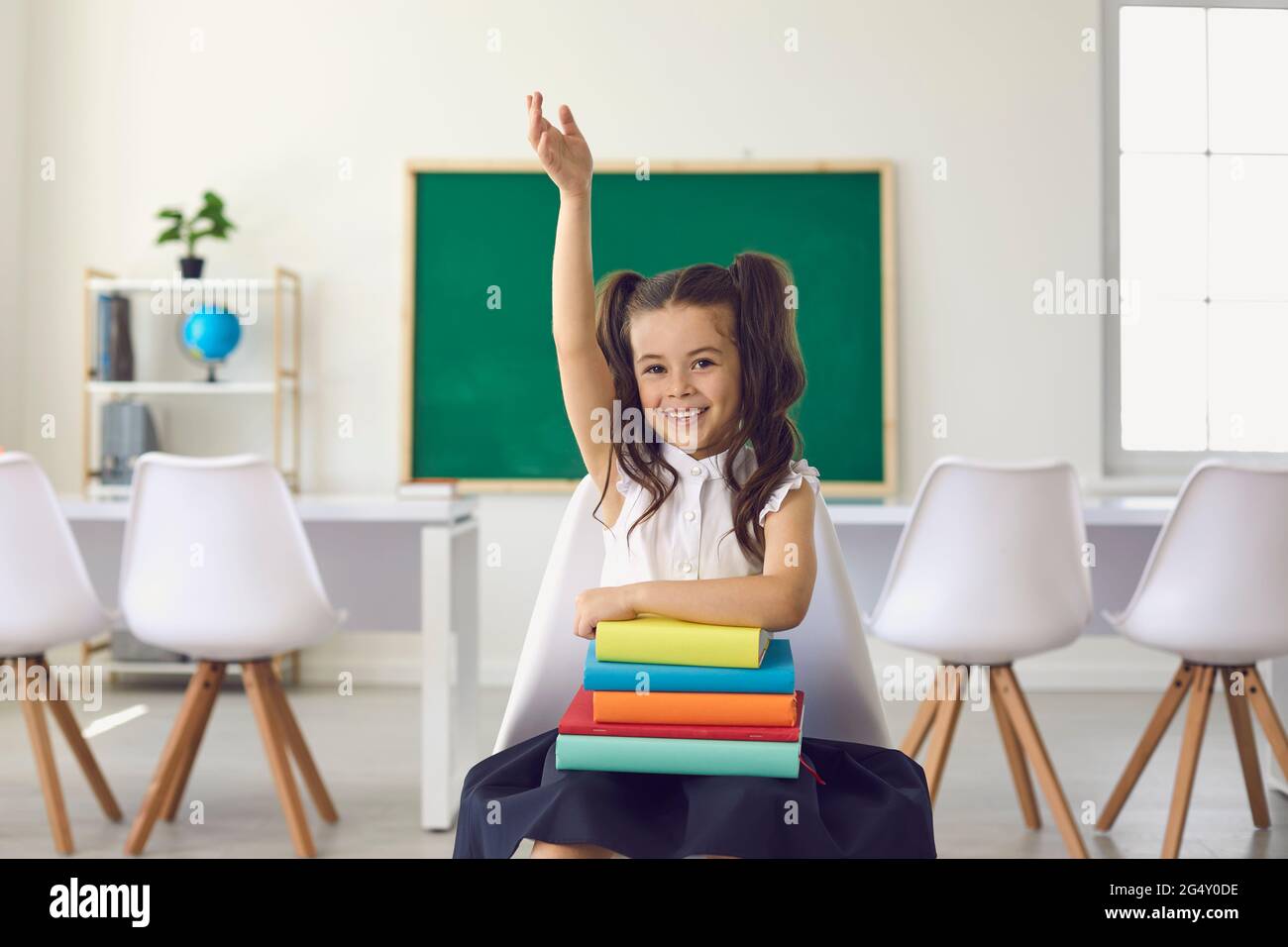 Concept de retour à l'école. Une petite écolière avec des livres dans ses mains a soulevé sa main contre le fond dans la classe à l'école primaire. Banque D'Images