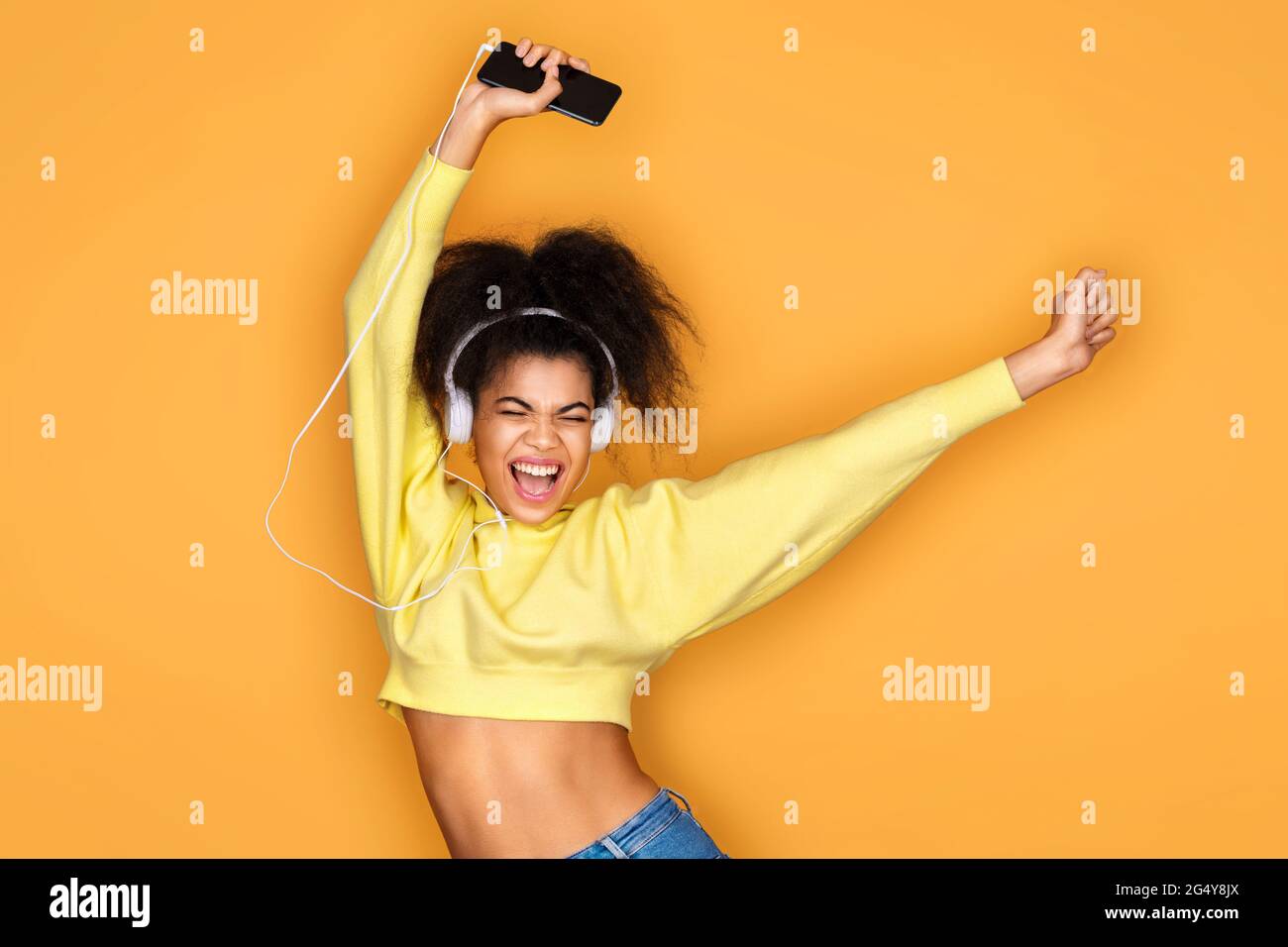 Joyeuse fille écoute la chanson préférée dans les écouteurs, lève la main dans la danse bouge et chante fort sur fond jaune Banque D'Images