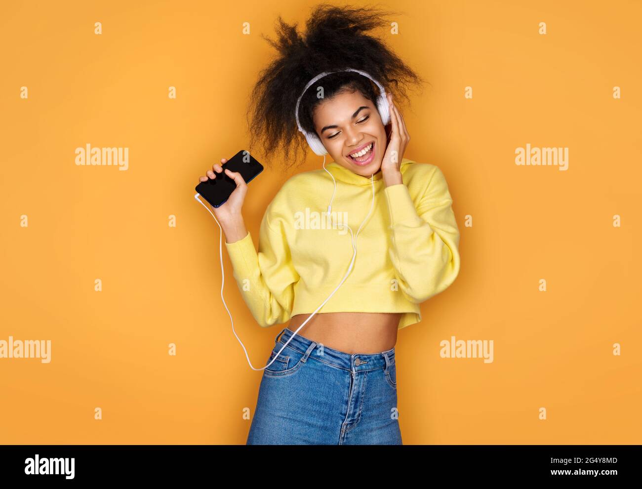 Une jeune fille écoute de la musique pour se détendre, tient son smartphone et porte un casque sur fond jaune Banque D'Images