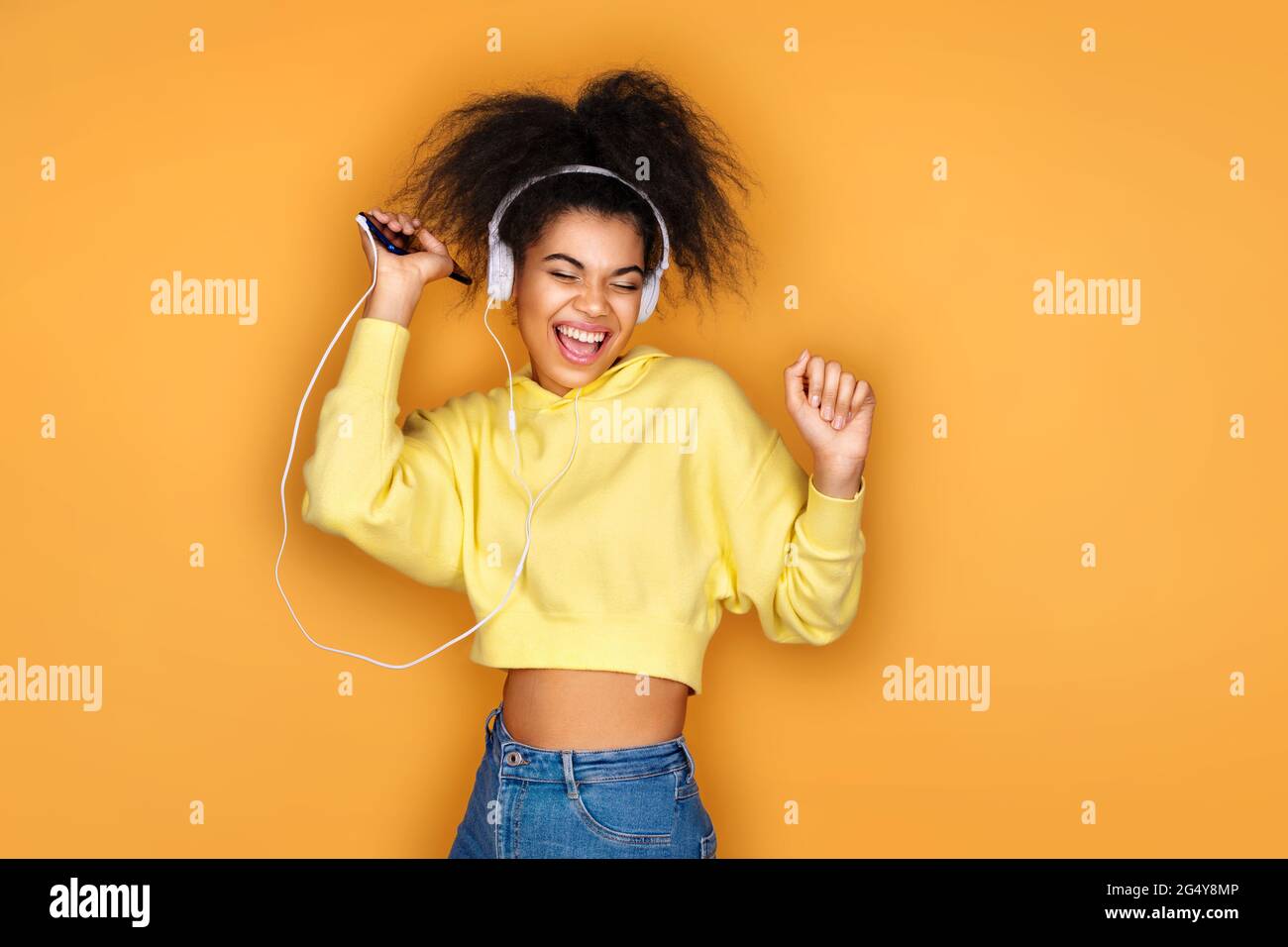 Joyeuse fille utilise des écouteurs et un smartphone, écoute de la musique, chante des chansons à haute voix sur fond jaune Banque D'Images