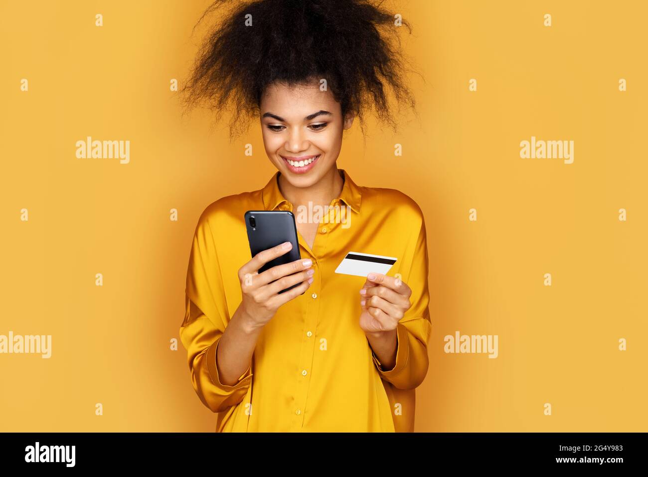 Jeune fille fait un paiement, en utilisant une carte de crédit et un smartphone. Photo d'une fille afro-américaine sur fond jaune Banque D'Images
