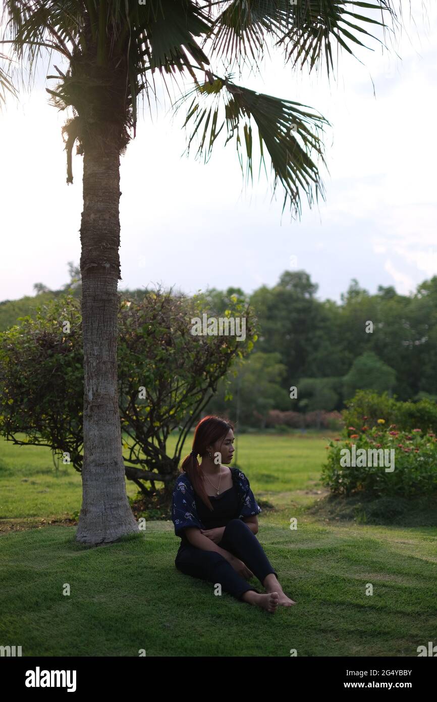 Une femme thaïlandaise est assise sous un palmier sur une pelouse taillée, donnant sur un parc pendant l'heure d'or Banque D'Images