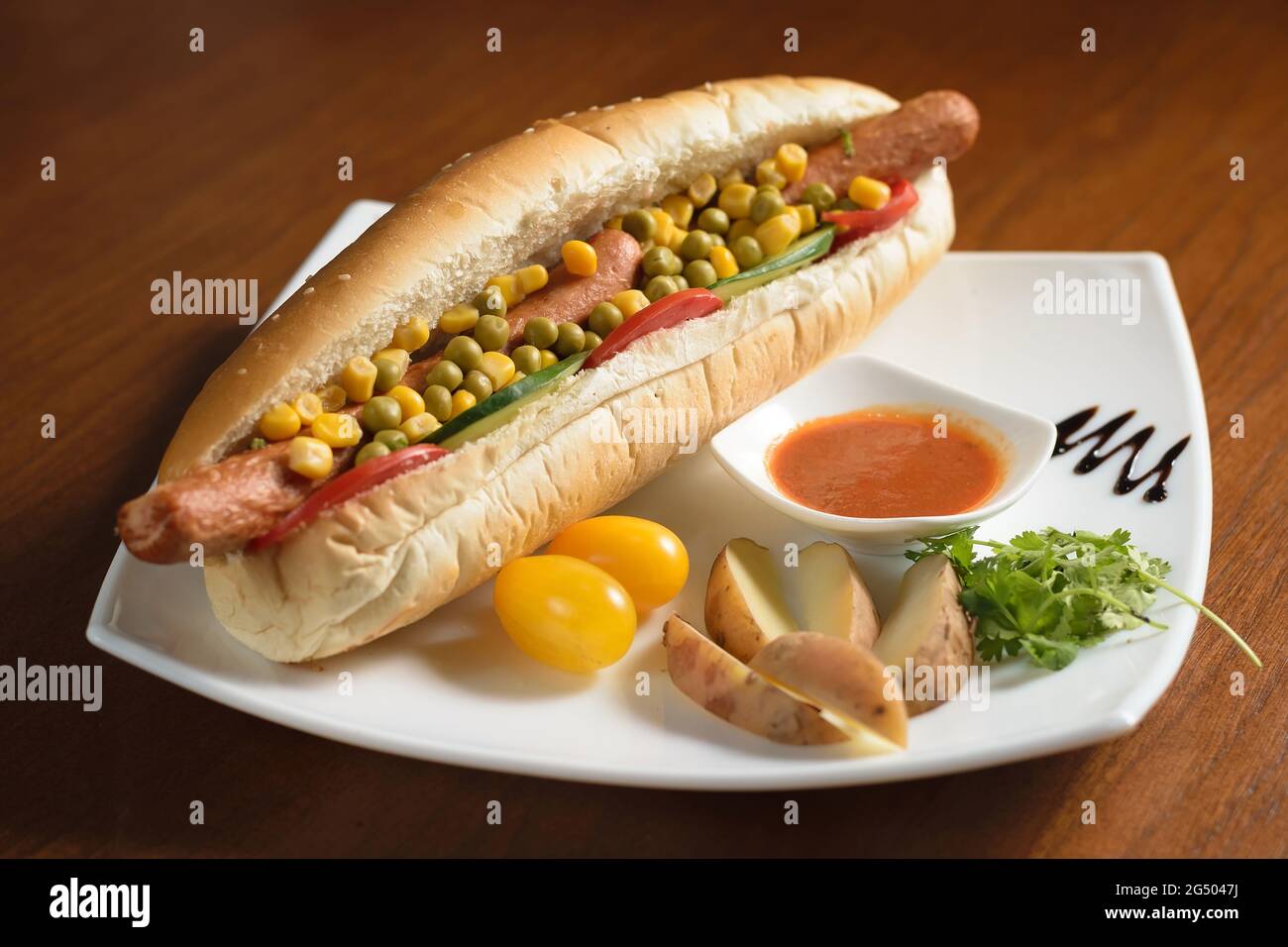 Long hot dog avec deux saucisses, petits pois verts et maïs, avec des tranches de tomates et de concombres, sur une assiette blanche avec sauce, pommes de terre et toma de cerise Banque D'Images