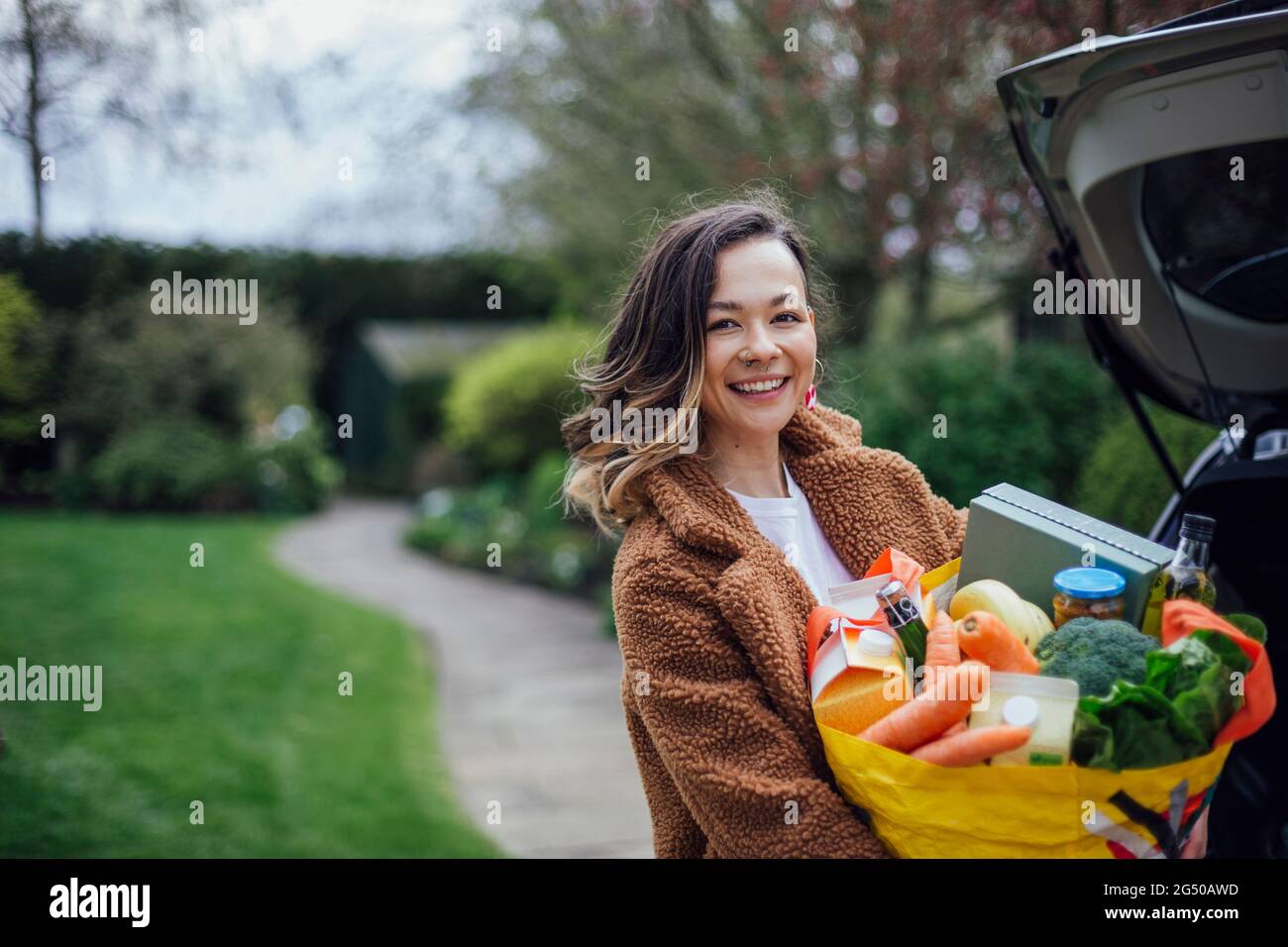 Une jeune femme souriant, regardant l'appareil photo et tenant un sac de transport réutilisable rempli de provisions. Elle déchargeant la voiture après avoir fait des achats de nourriture. Banque D'Images