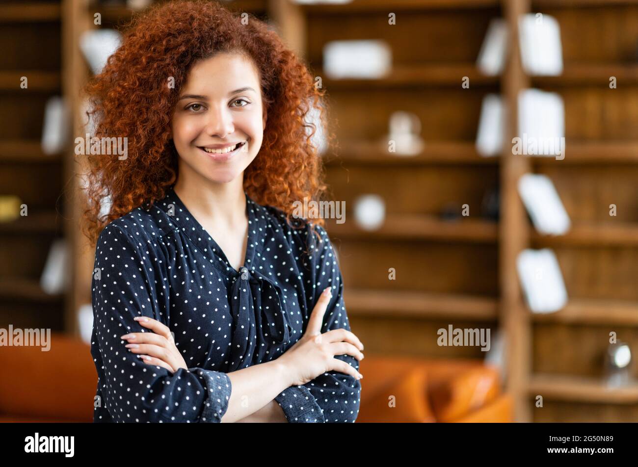 Femme ambitieuse, souriante et murée, debout dans l'espace de bureau avec ses bras croisés et regardant la caméra, charmante étudiante aux cheveux rouges dans la bibliothèque, femme entrepreneure prospère à l'intérieur Banque D'Images