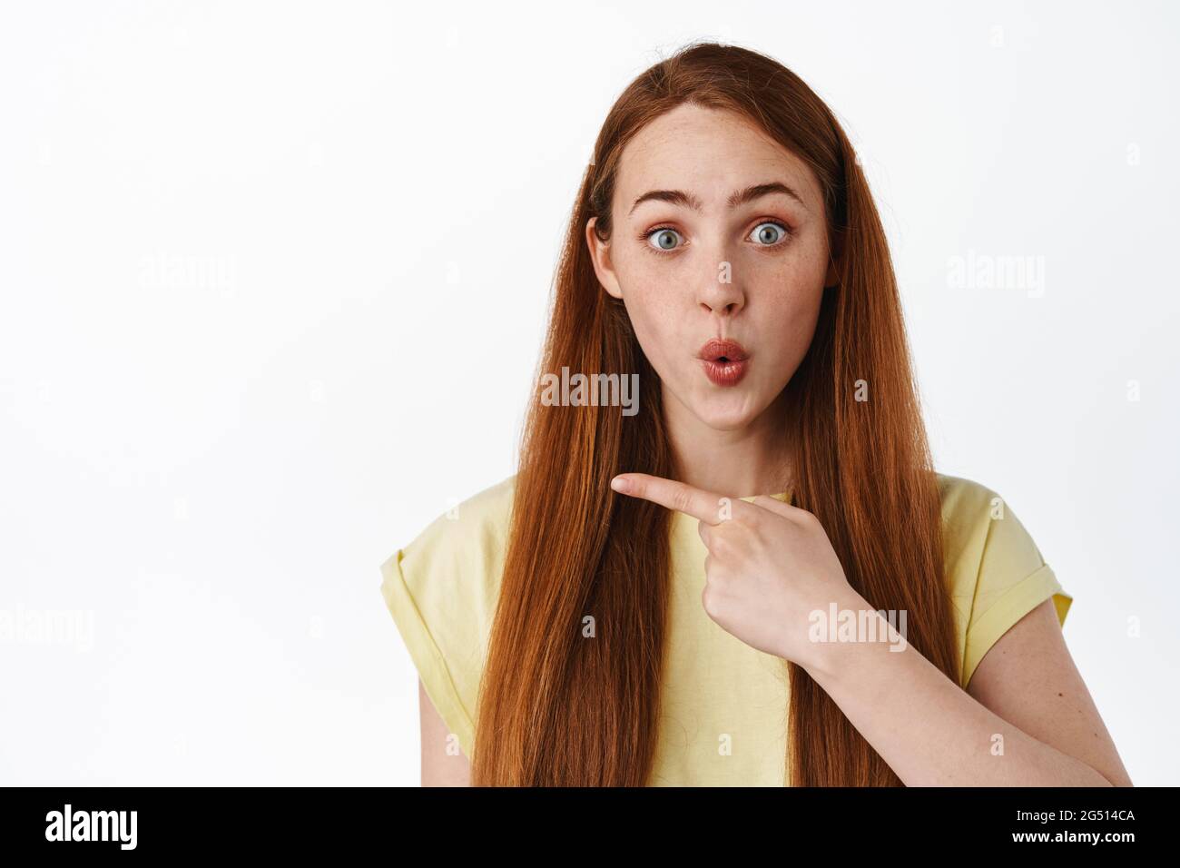 Gros plan de la femme de redhead amusé s'est curieuse avec la publicité, pointant vers la gauche et de dire wow impressionné, montrant la promotion, fond blanc Banque D'Images