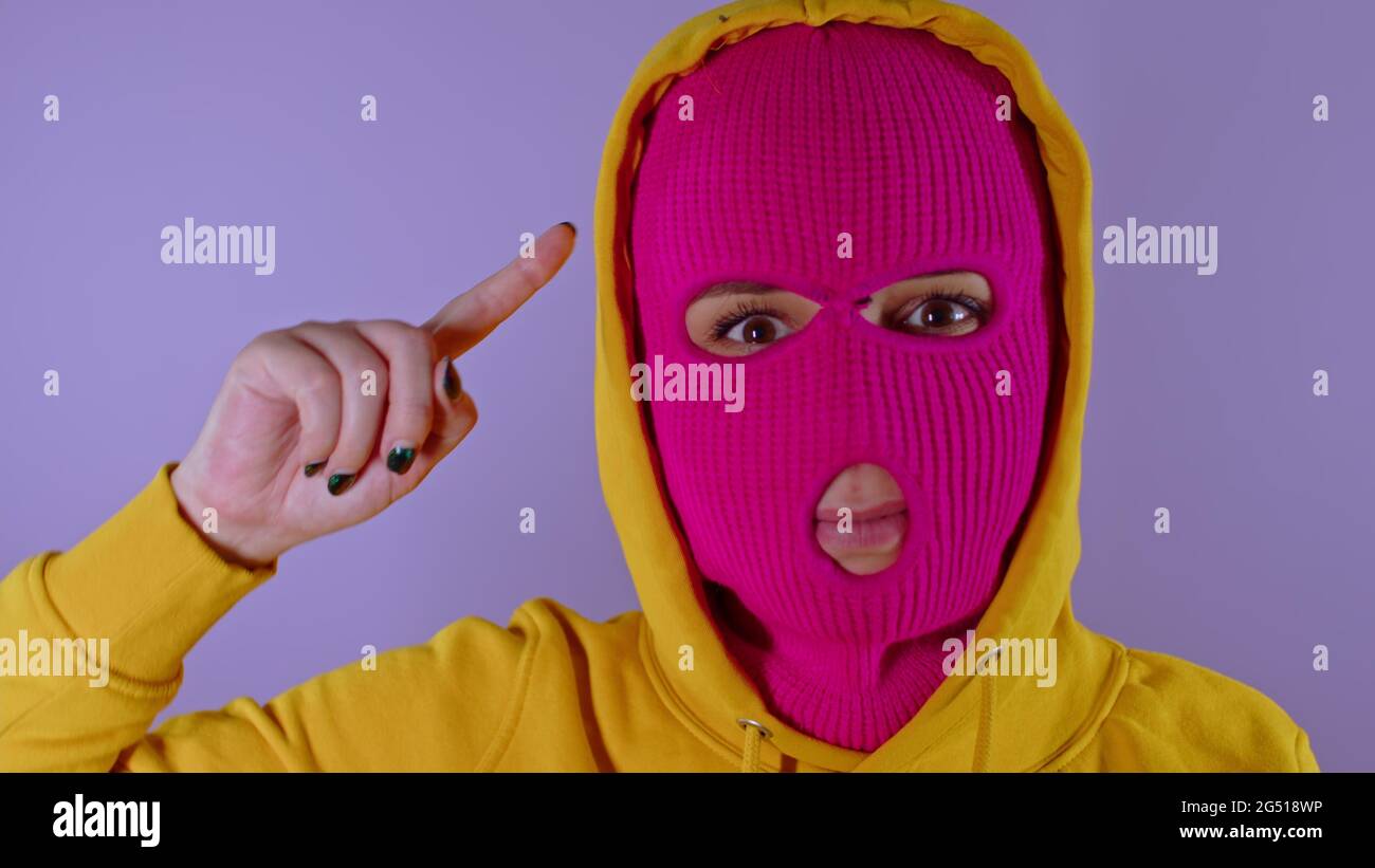 Gros plan de la jeune femme en balaclava rose et capot jaune avec un doigt de torsion sur son temple sur fond violet. Femelle surprise dans le masque Twists index fi Banque D'Images