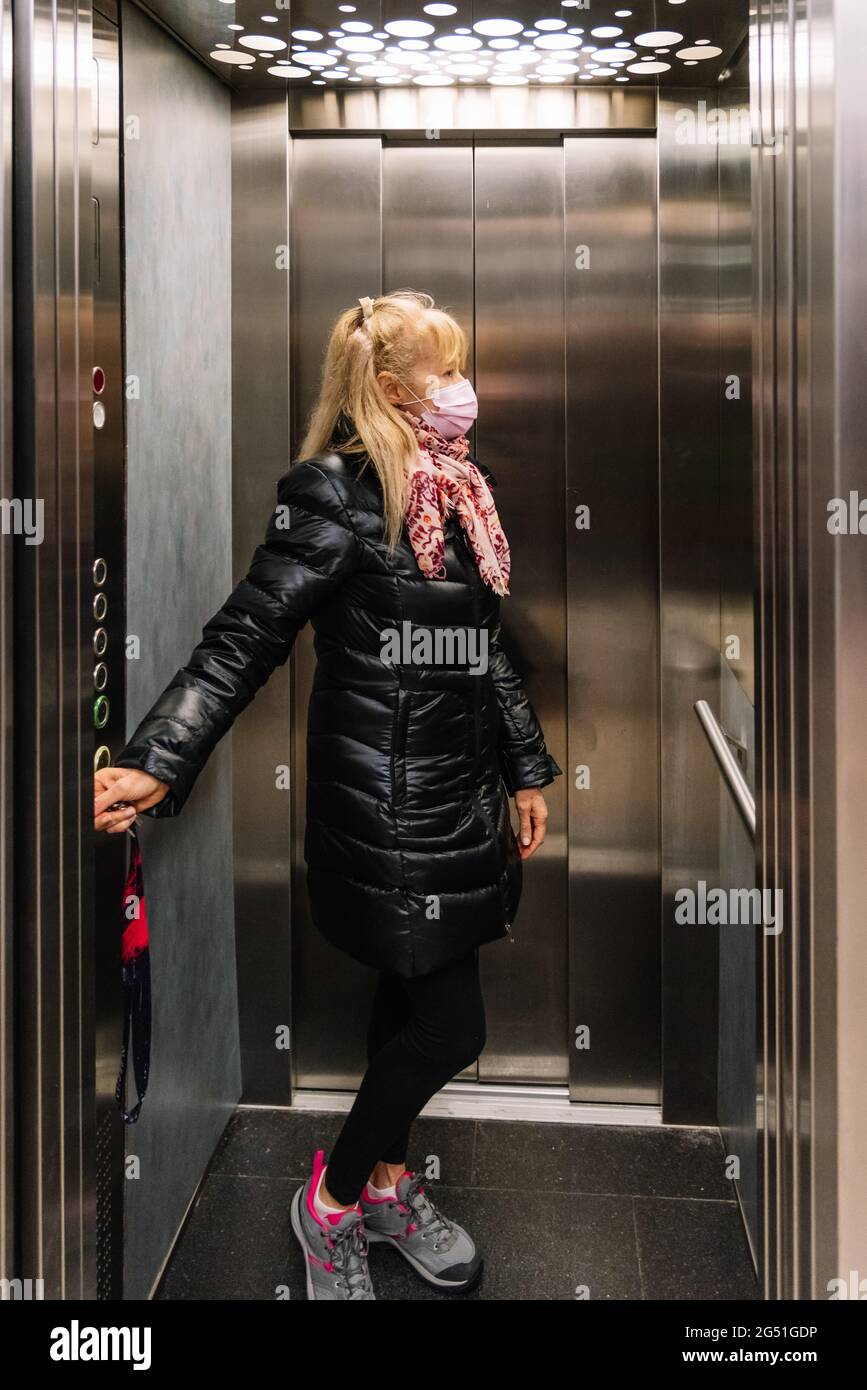 Femme blonde adulte portant un masque en appuyant sur les boutons d'un ascenseur pour rentrer à la maison. Banque D'Images