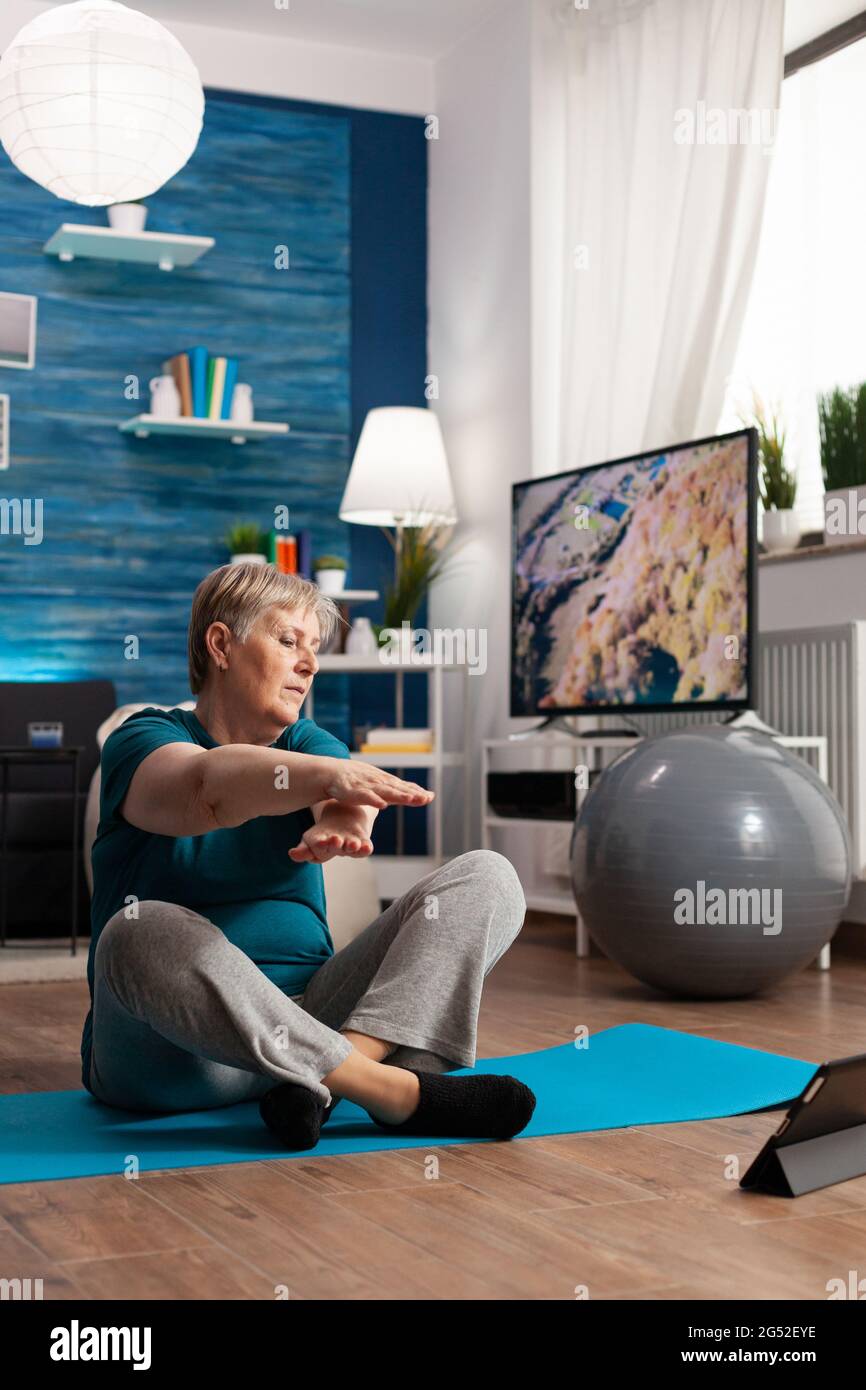 Femme sénior à la retraite dans des vêtements de sport assise sur un tapis de yoga regardant la leçon de fitness sur un ordinateur portable pratiquant l'exercice de bras amincissant le poids. Retraité qui travaille à la flexibilité du corps pendant l'entraînement de bien-être Banque D'Images