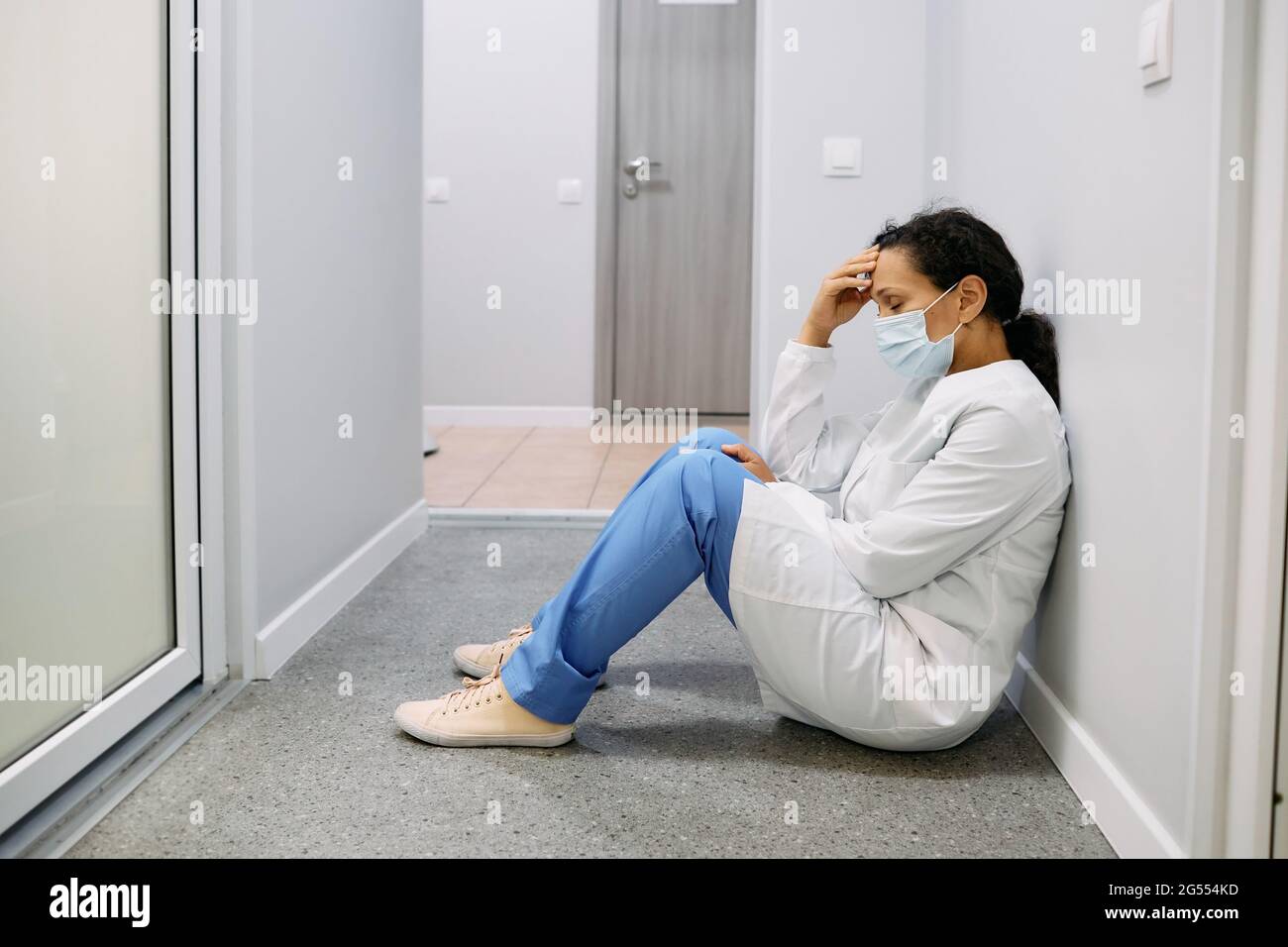 Une femme fatiguée médecin après une dure journée de travail dans une clinique médicale s'assoit au sol dans un hôpital de couloir avec ses yeux fermés, portant un masque médical. Pand Banque D'Images
