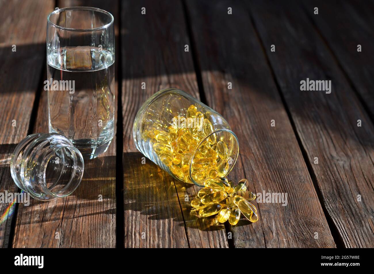 Supplément diététique en capsules de gélatine jaune dans un pot en verre à côté d'un verre d'eau sur une table en bois. Vitamines pour la santé et la longévité. Banque D'Images