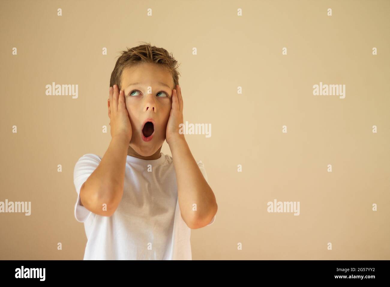Un garçon surpris ou effrayé de 7-10 ans dans un T-shirt blanc se tient et crie avec ses mains sur ses joues sur un fond beige. Copier l'espace. Banque D'Images