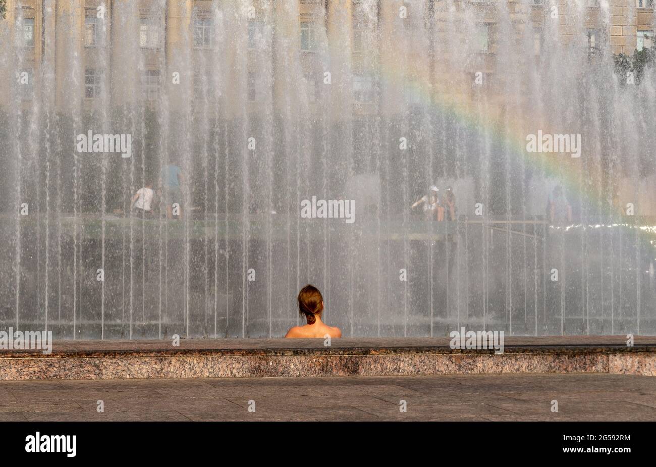 La tête et les épaules d'une femme, vue du dos, contre la fontaine arrosée, avec arc-en-ciel, par une chaude journée d'été Banque D'Images