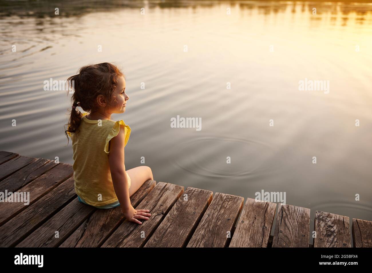 Une belle petite fille s'assoit sur une jetée en bois au bord du lac et  profite de la chaleur d'une soirée d'été. La belle lumière du coucher du  soleil se reflète dans