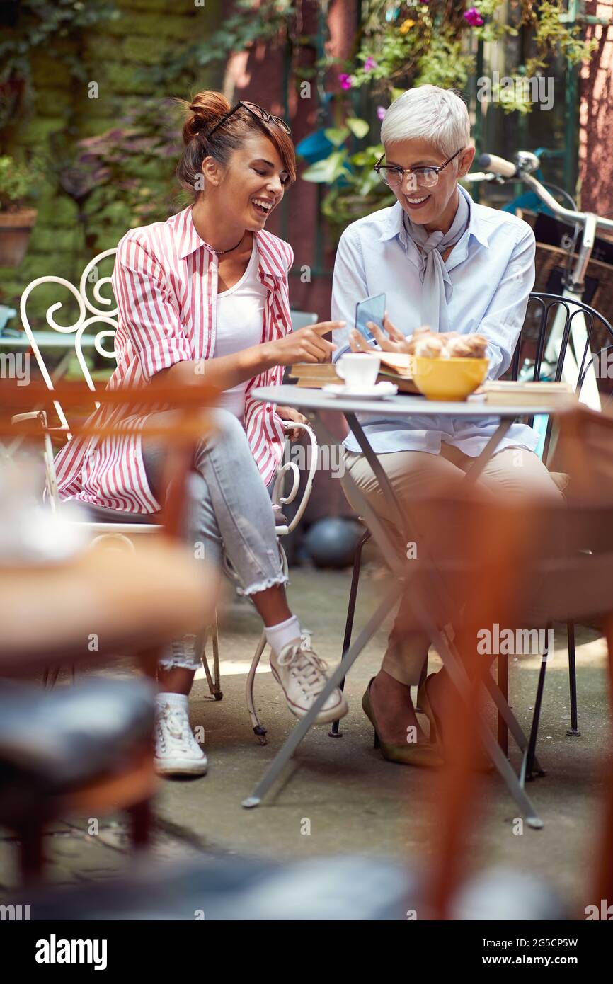 deux femmes, âgées et jeunes, assis dans un café extérieur, parlant, souriant, regardant le téléphone cellulaire transparent. technologie de nouvelle génération. Banque D'Images