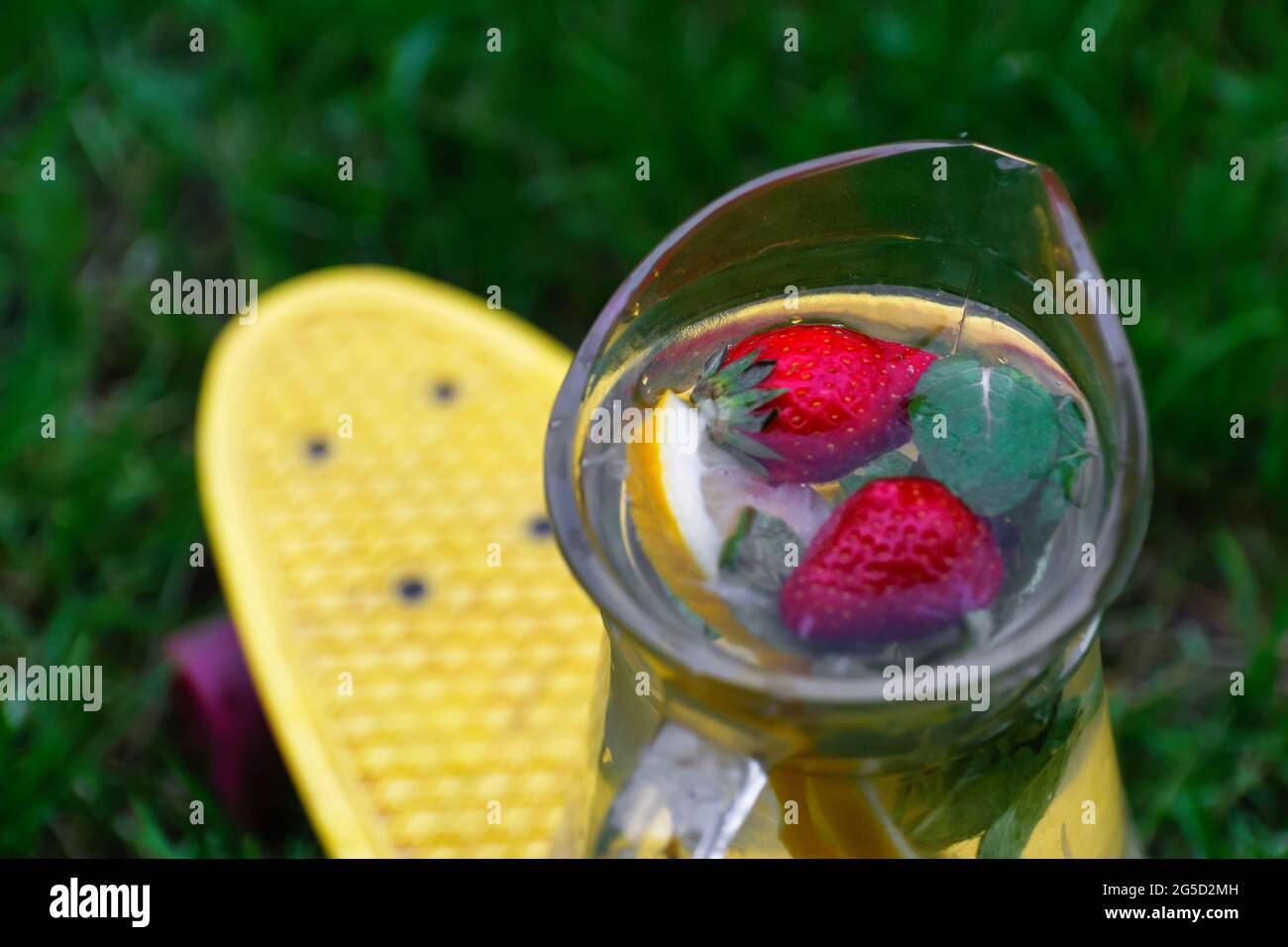 Flou artistique en verre de la limonade à la fraise, trancher le citron et les feuilles de menthe sur le panneau jaune de penny, planche à roulettes. Arrière-plan d'herbe flou. Pichet de Banque D'Images