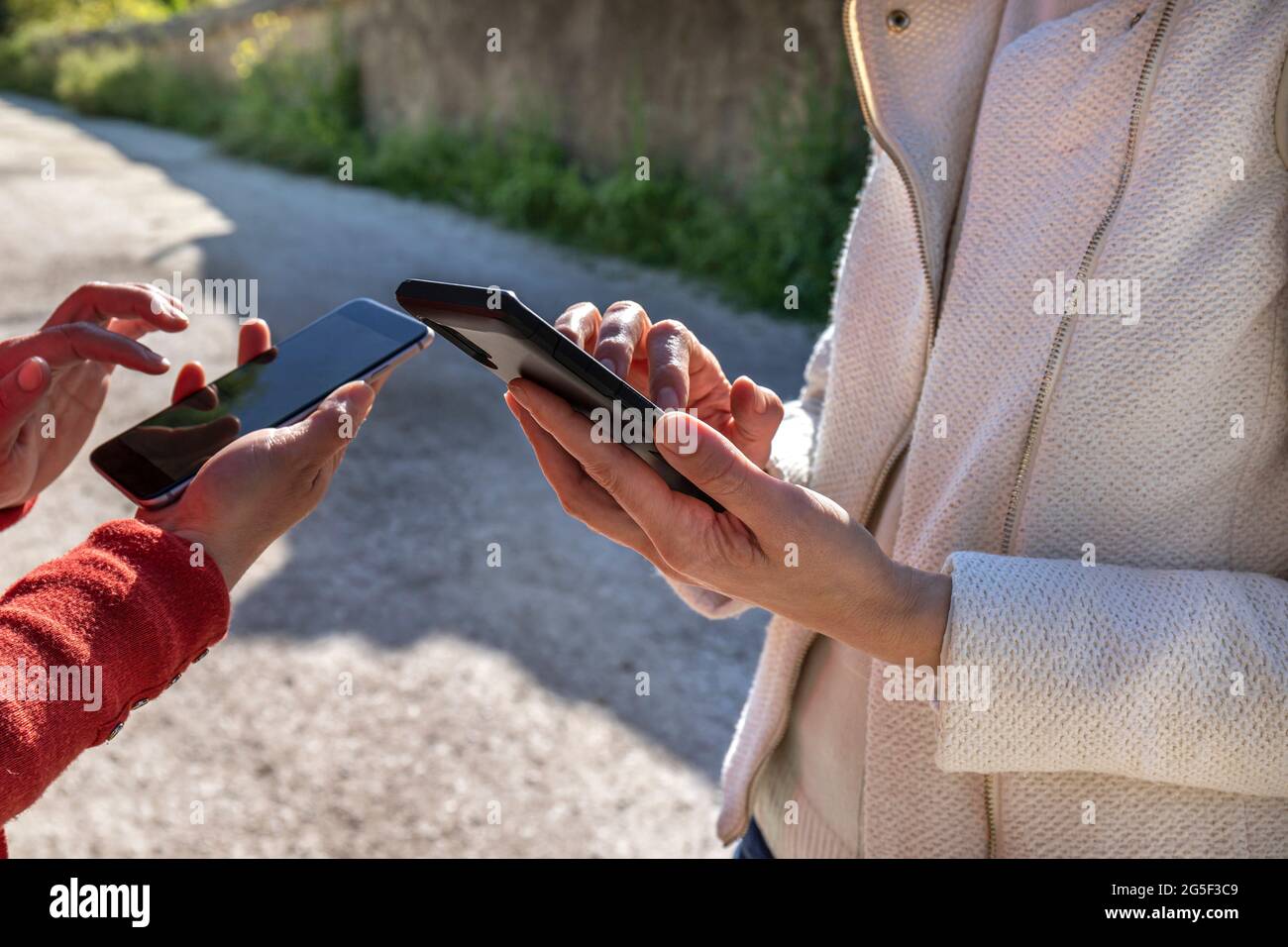 Gros plan de deux mains de filles utilisant leur smartphone dans un parc. Une fille porte une veste blanche et l'autre un chandail rouge. Banque D'Images