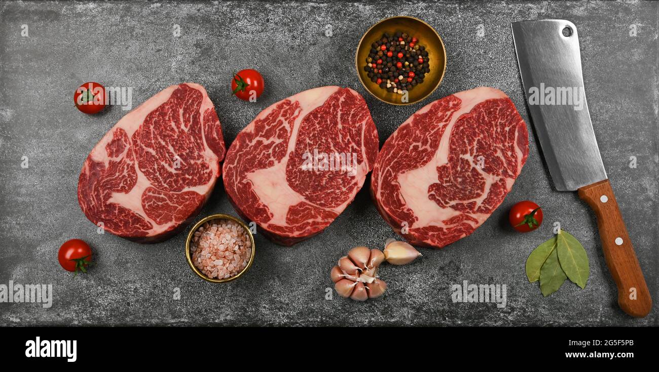 Gros plan sur trois steaks de bœuf de bœuf brut de choix marbré avec couteau à laver et épices, sur fond de table en pierre, vue en hauteur, juste au-dessus Banque D'Images