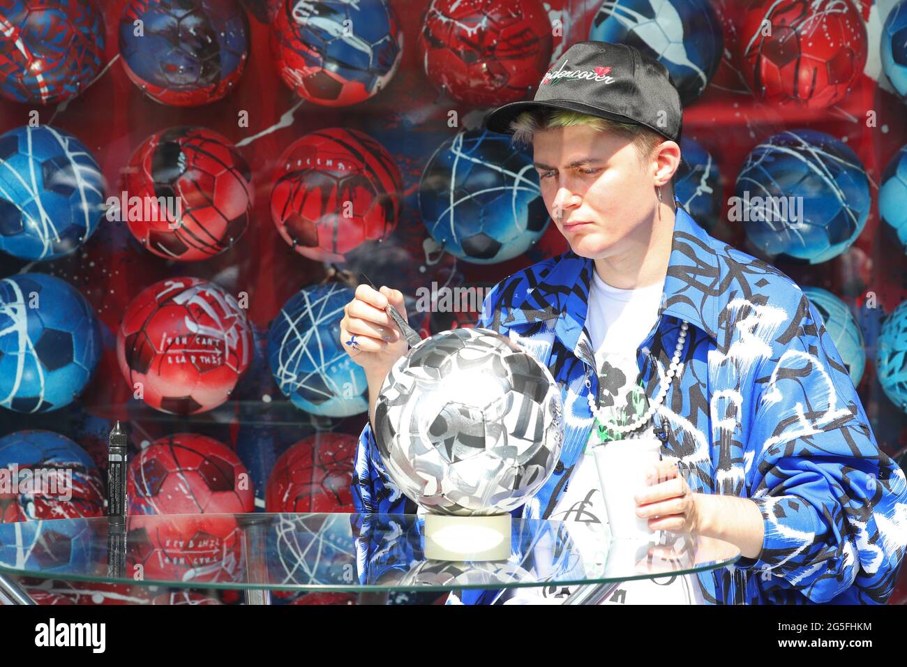 Saint-Pétersbourg, Russie. 27 juin 2021. L'artiste calligraphique Arseny  Pyzhenkov aka Pokras Lampas peint une balle de football à côté de son  installation artistique installée dans le village de football de la place