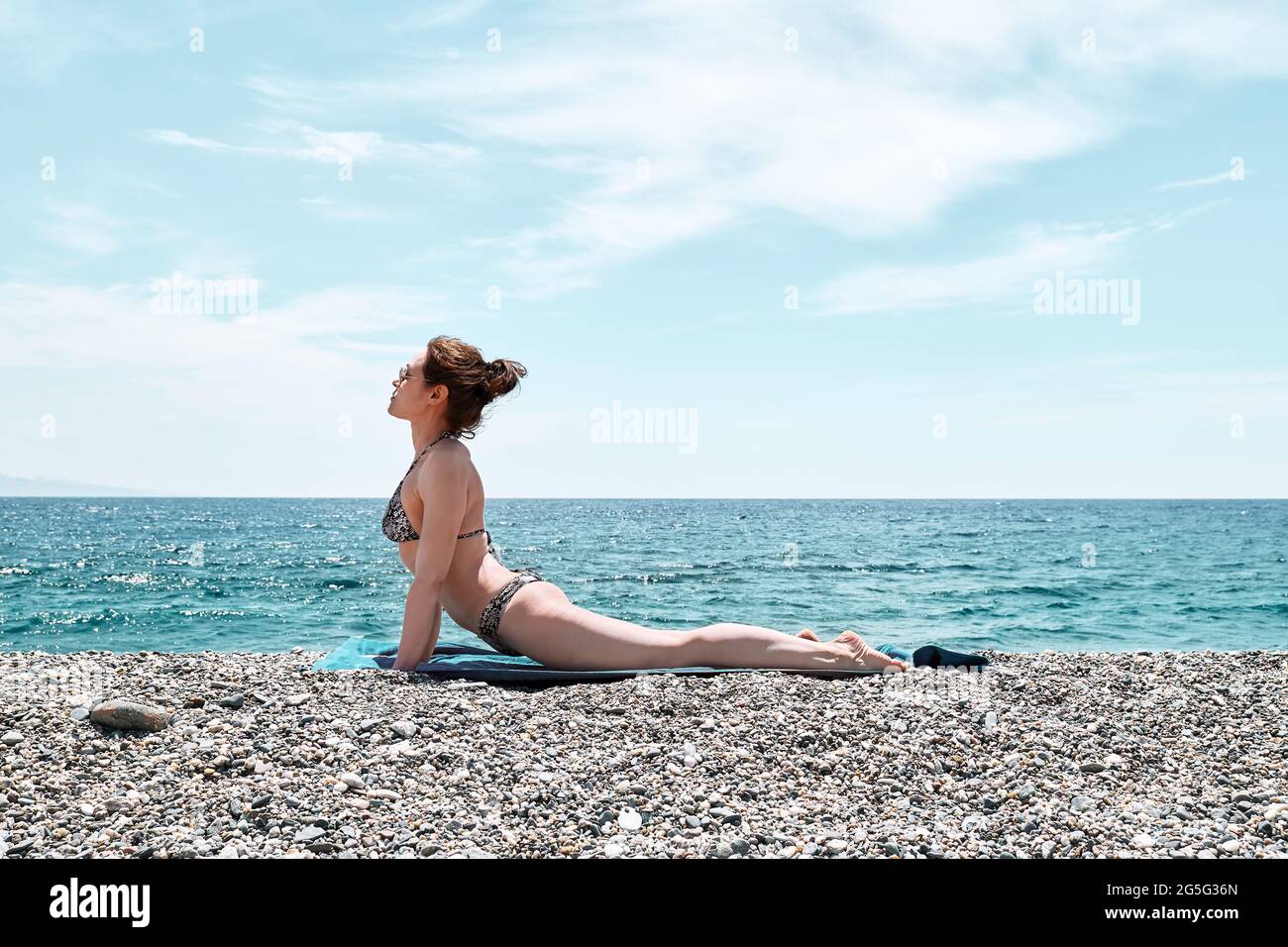 Femme mince faisant du yoga sur la plage avec la mer bleue en arrière-plan. Renforcement des muscles des jambes et coordination du corps. Pratiquer le yoga en plein air. Banque D'Images