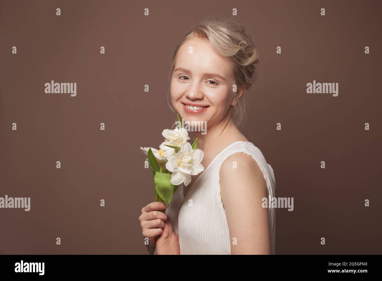 La beauté blonde dans le maquillage léger souriant et tenant des fleurs Banque D'Images