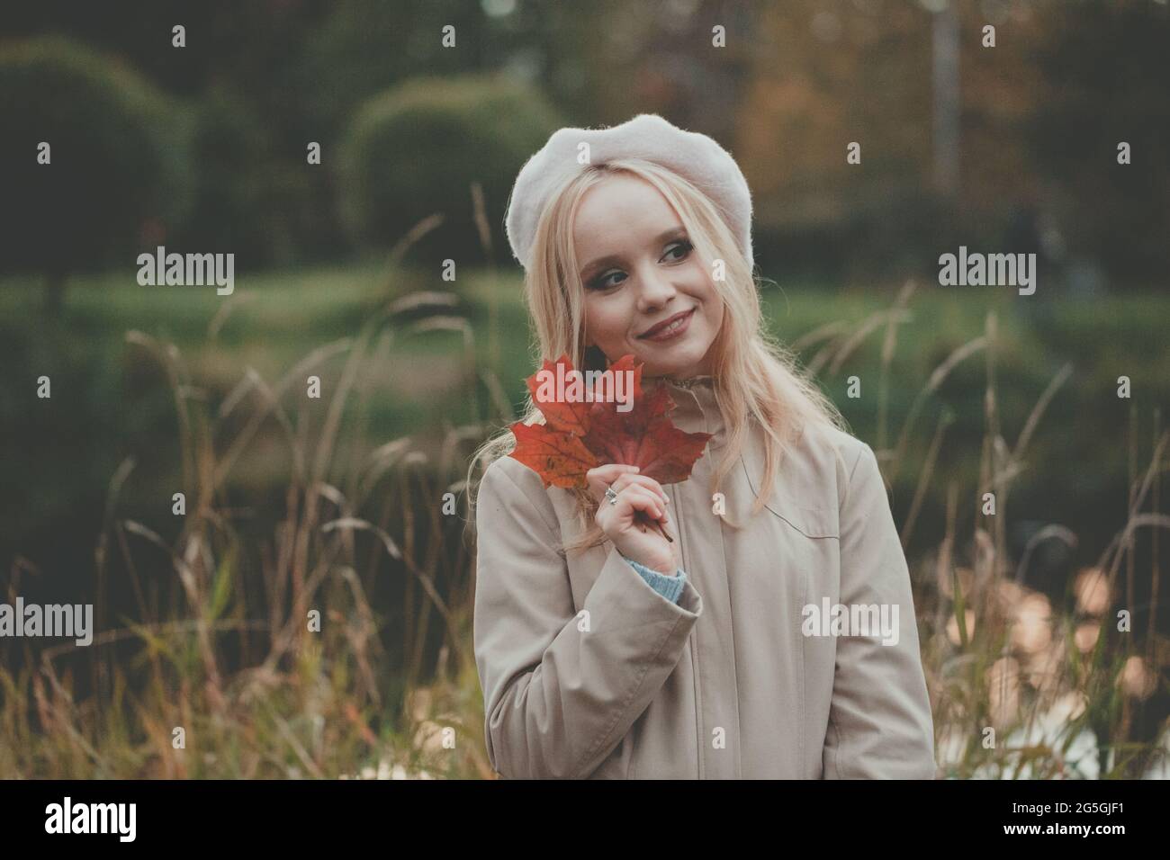 Portrait extérieur de jeune belle heureuse femme souriante avec de longs cheveux portant un béret beige et un manteau tenant des feuilles d'automne rouges dans le parc d'automne Banque D'Images