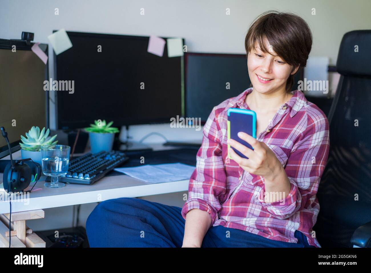 Femme souriante regardant l'écran du téléphone tout en étant assise sur son bureau ou son lieu de travail. Faites une pause. Lire la bonne nouvelle. Travail à distance Banque D'Images