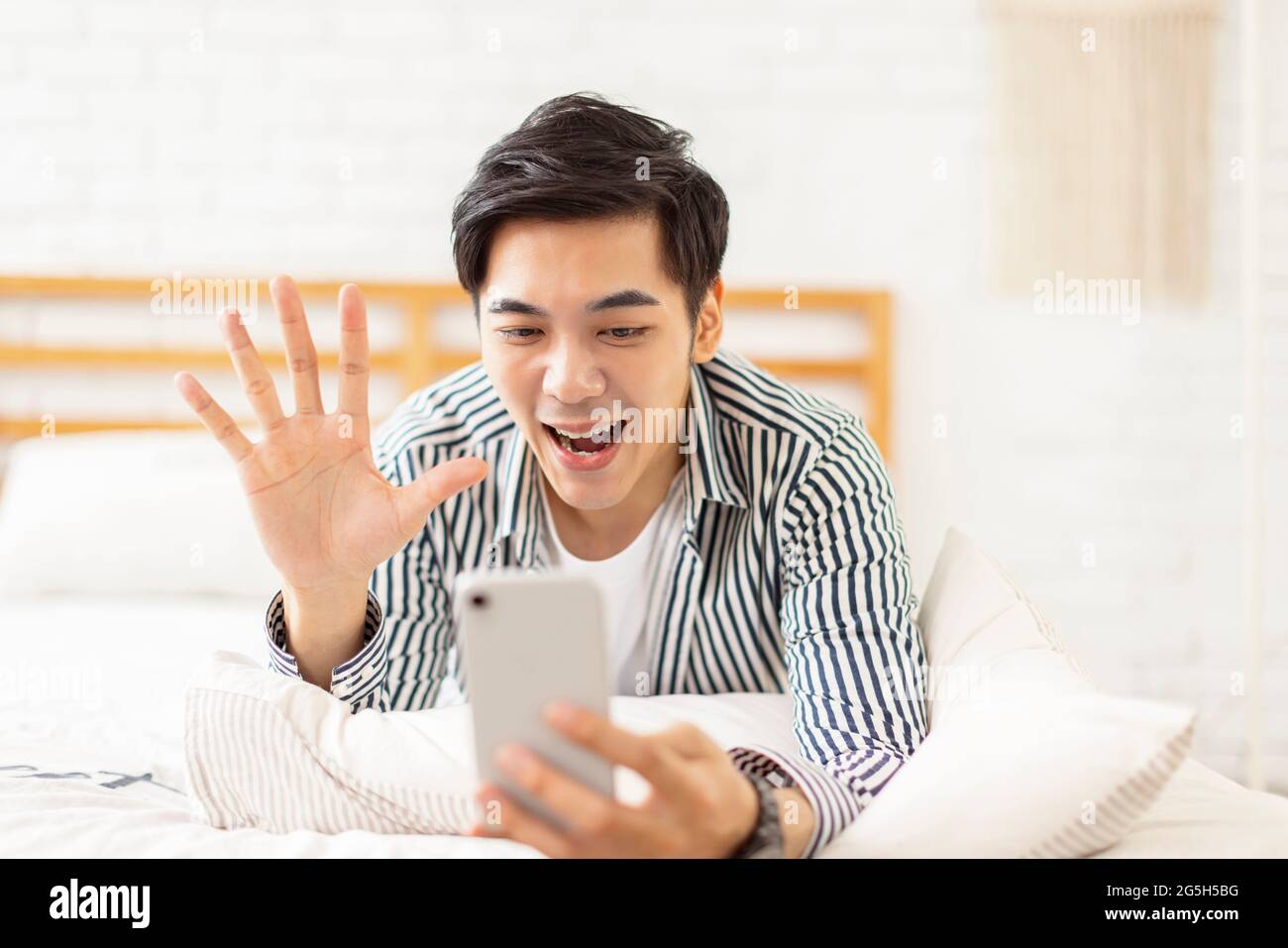 Beau jeune homme allongé sur le lit, utilisant un téléphone portable pour faire un appel vidéo, et en agitant à l'écran pour se saluer Banque D'Images