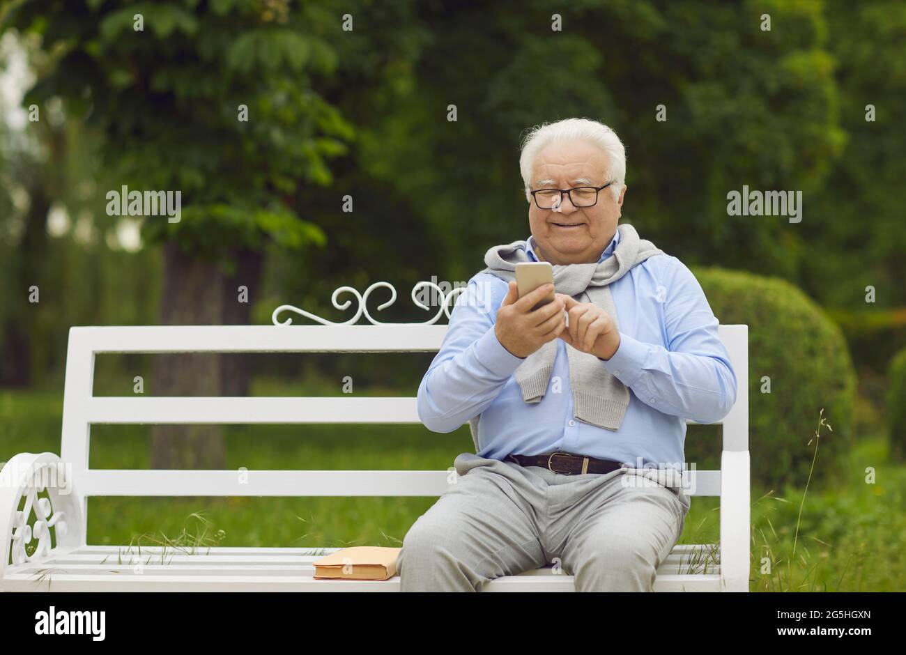 Homme âgé heureux utilisant un téléphone portable tout en étant assis sur un banc dans un parc d'été vert Banque D'Images