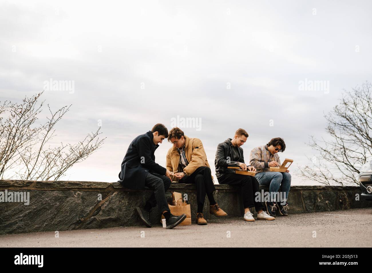 Amis masculins multi-ethniques ayant de la nourriture tout en étant assis sur le mur de soutènement contre le ciel Banque D'Images