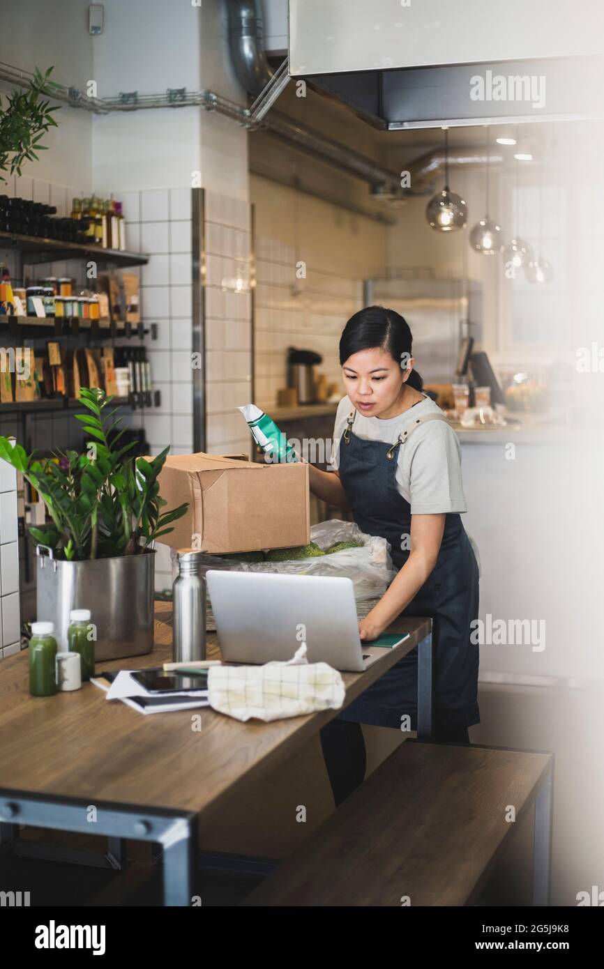 Femme propriétaire travaillant sur un ordinateur portable tout en tenant le paquet de nourriture dans le magasin Banque D'Images