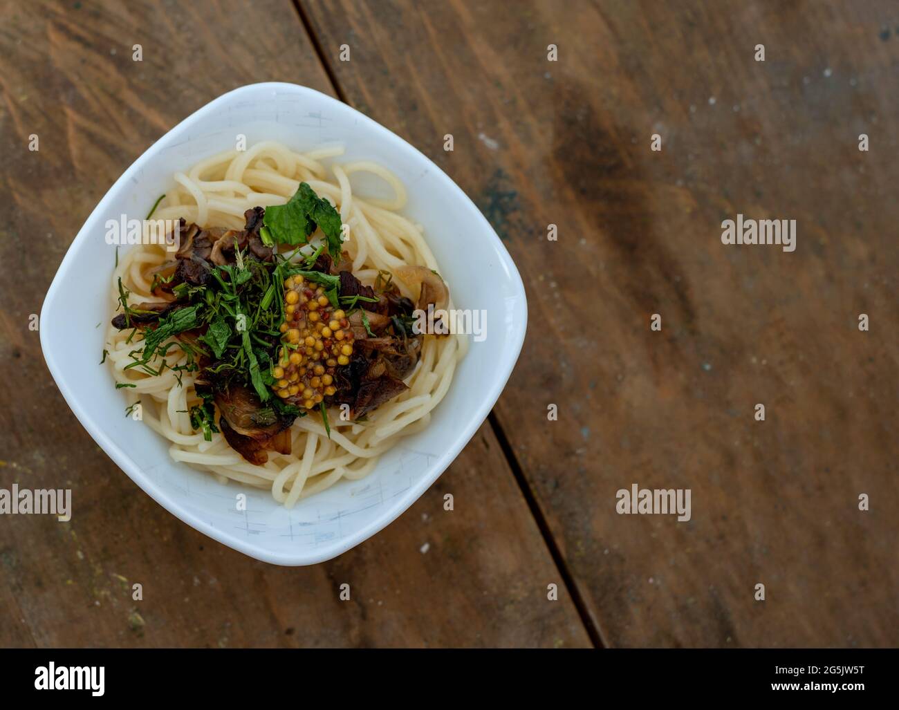 Spaghetti fraîchement cuits placés dans une petite assiette carrée en céramique avec un pot aux champignons parsemé d'herbes et de moutarde française sur fond de bois Banque D'Images