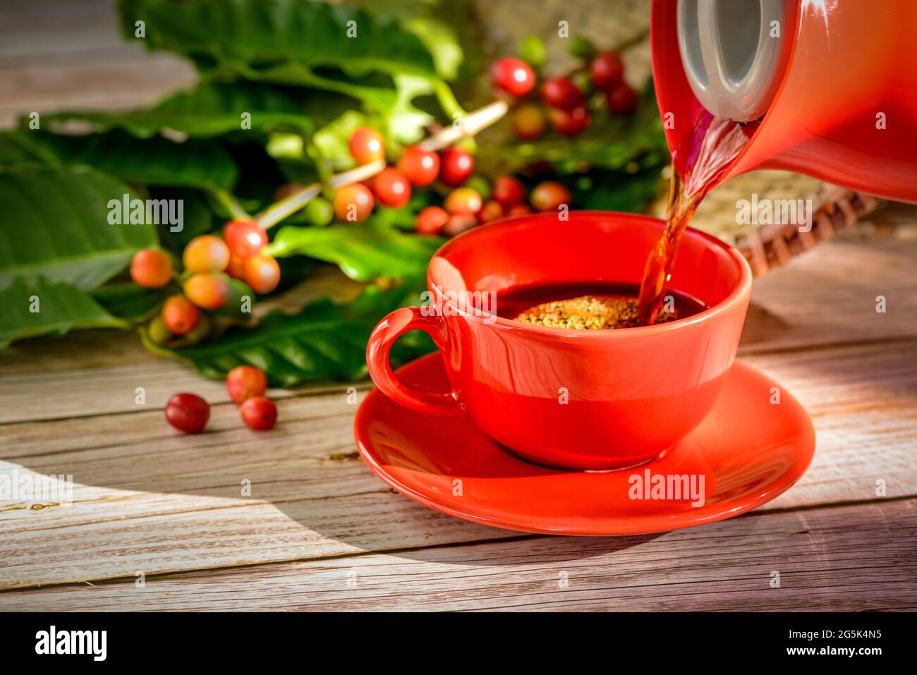 Le café est versé dans une tasse rouge avec des graines de café et des feuilles sur une table en bois. Banque D'Images