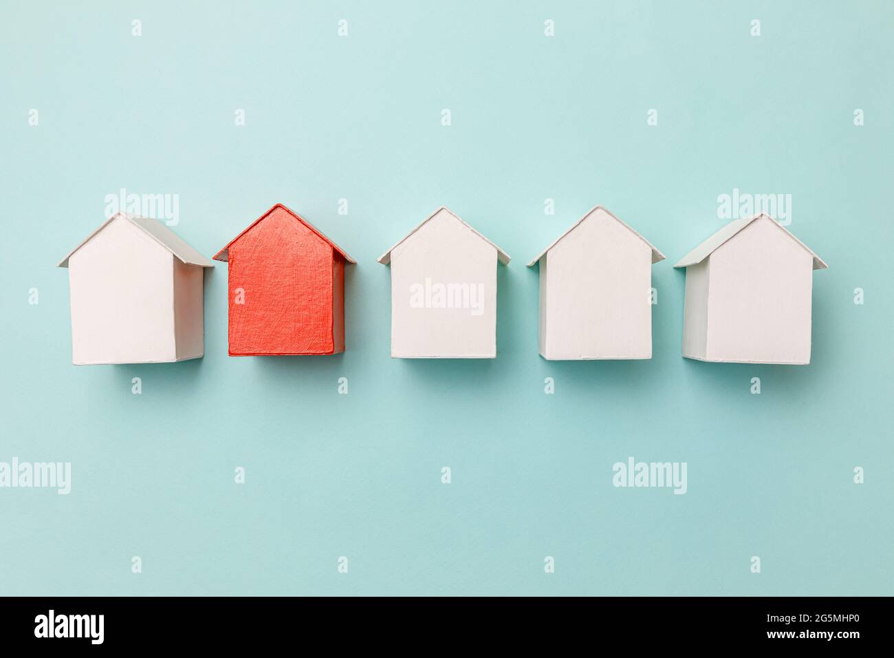 Conception simple de pose plate avec miniature de jouet rouge maison modèle parmi les maisons blanches isolées sur fond bleu pastel. Industrie de l'immobilier Banque D'Images