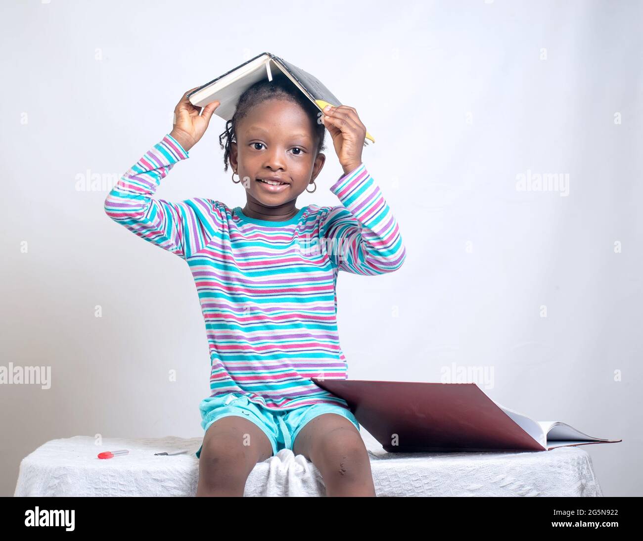 Adorable fille africaine avec le style de cheveux tissé place heureusement un livre sur sa tête tout en étudiant pour montrer comment intéressant l'éducation est devenue Banque D'Images