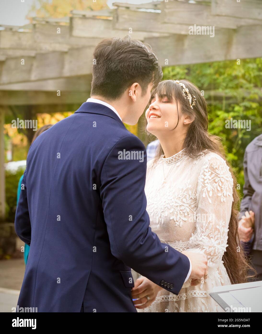 Mariée biraciale et marié se préparant à embrasser pendant la cérémonie de mariage à l'extérieur sous pergola Banque D'Images