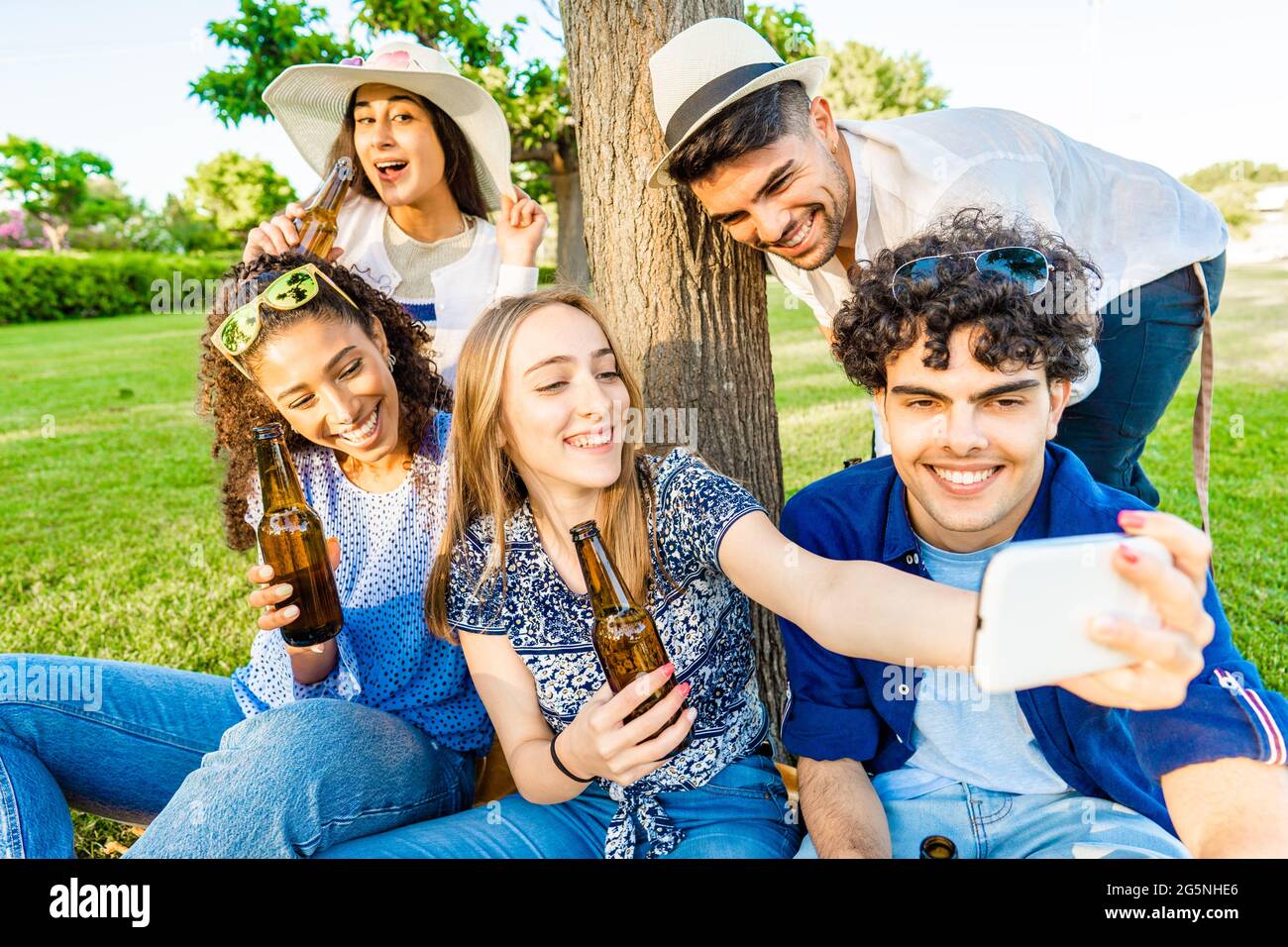 Cinq jeunes amis multiraciaux s'amusent ensemble dans un pique-nique au parc de la ville en buvant de la bière à la bouteille. Jeune femme blonde prenant un autoportrait avec Banque D'Images