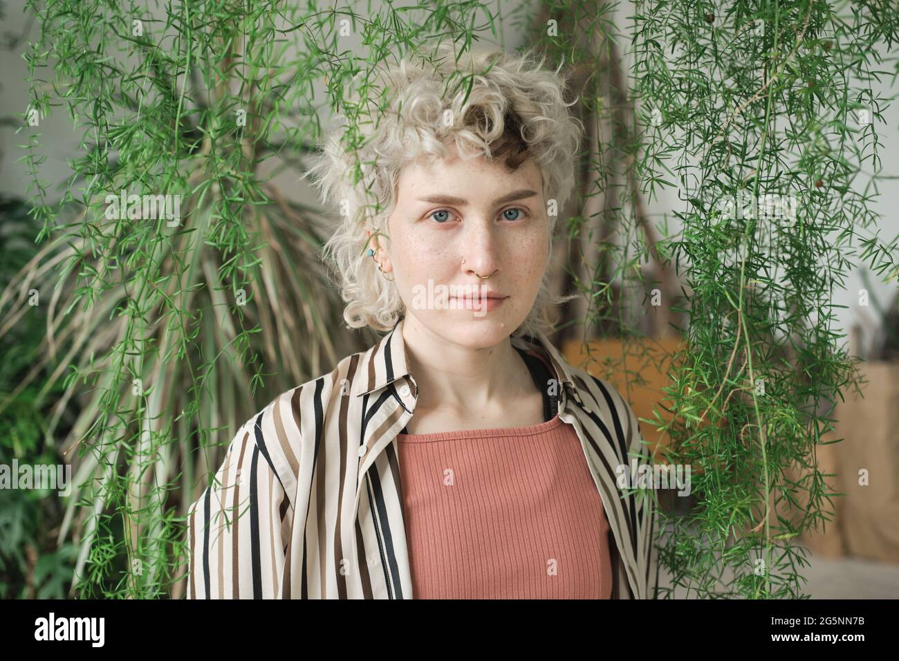 Portrait d'une jeune femme aux cheveux blonds et bouclés regardant un appareil photo debout parmi des plantes vertes Banque D'Images