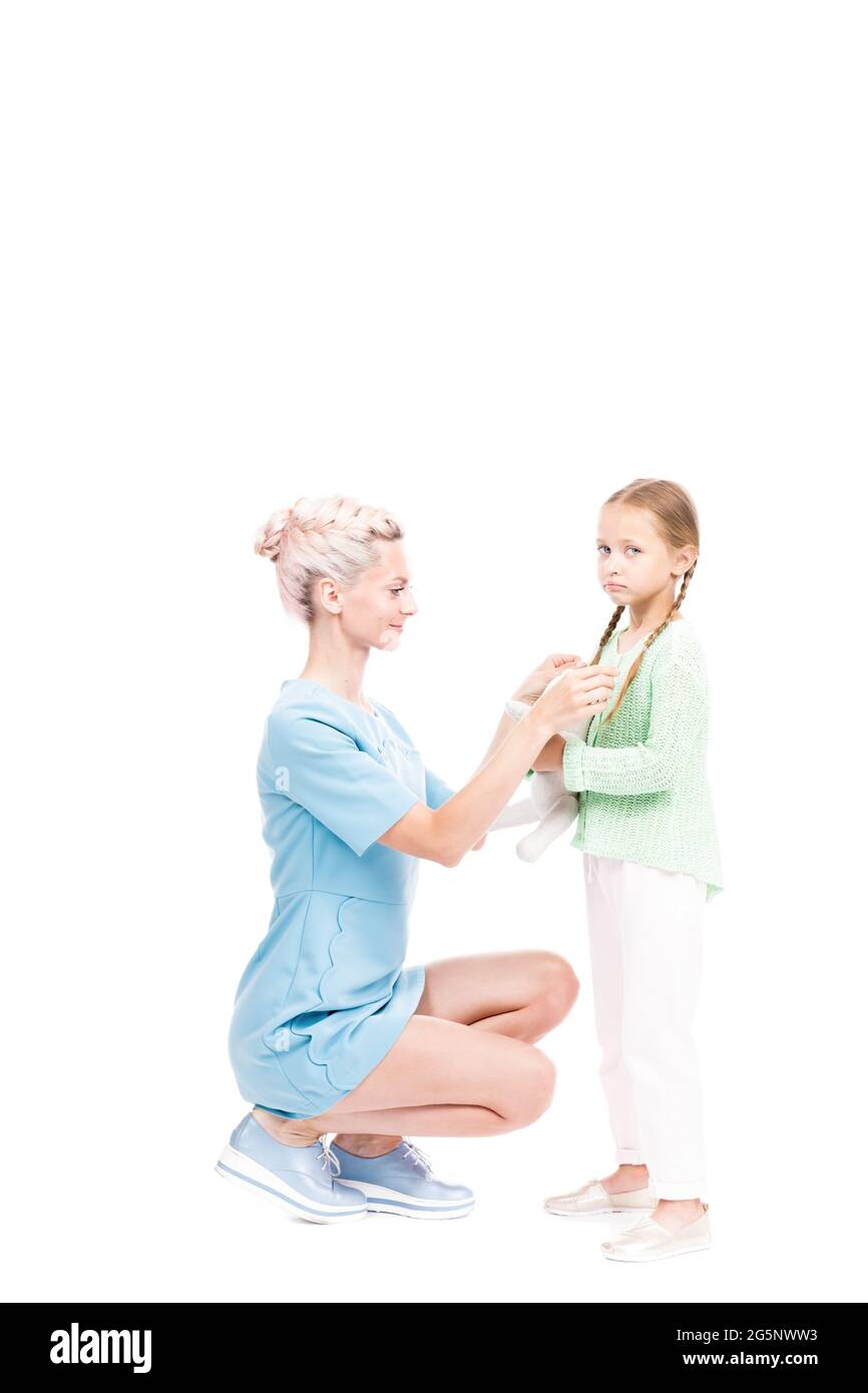 Vue verticale de la mère moderne, en longueur, assise devant sa fille, fixant ses cheveux, fond blanc Banque D'Images