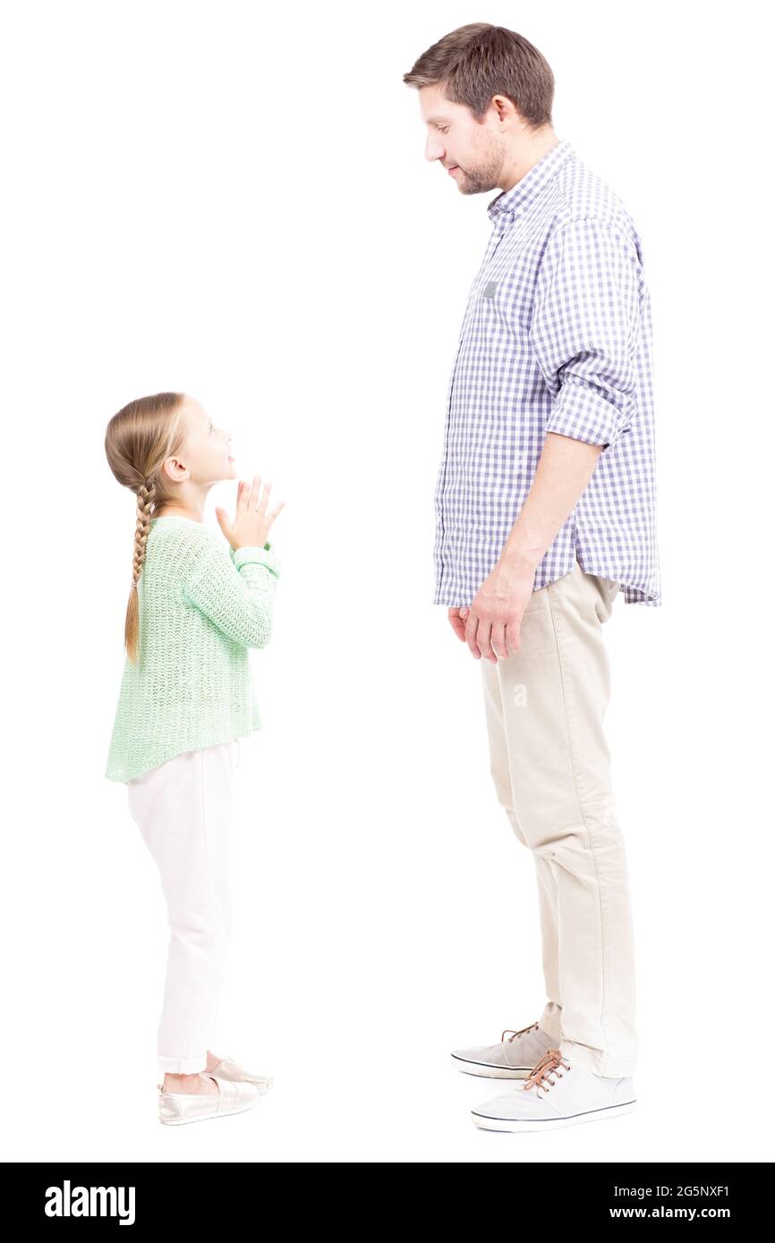 Vue verticale pleine longueur isolée de petite fille mignonne debout devant son père lui supplier quelque chose, fond blanc Banque D'Images