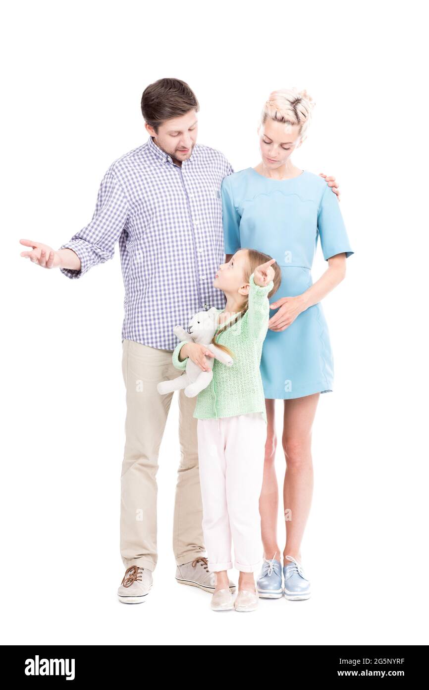 Photo verticale pleine longueur de petite fille caucasienne montrant quelque chose à ses parents pointant le doigt vers l'appareil photo, fond blanc Banque D'Images