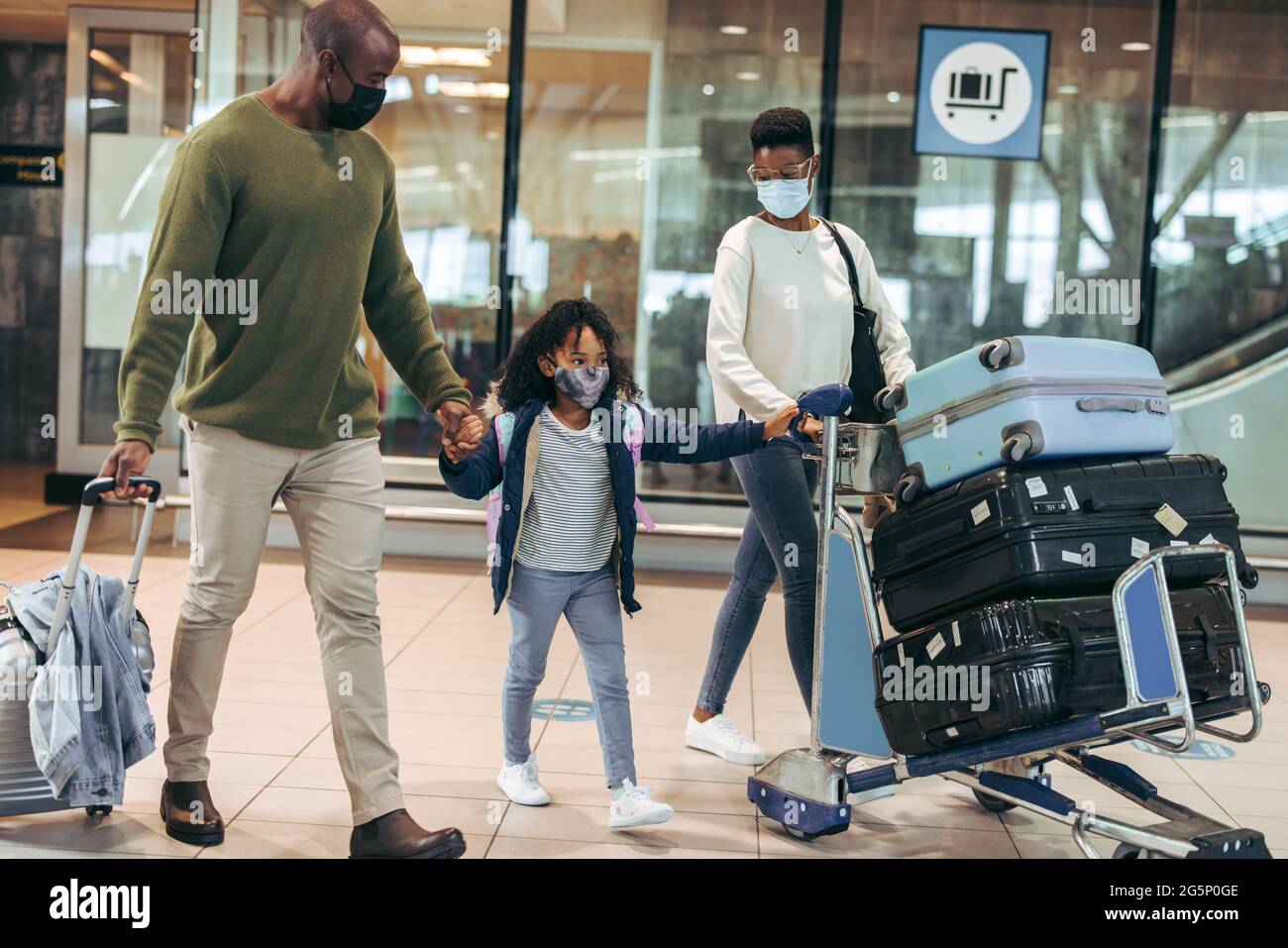 Homme et femme avec une jeune fille voyageant pendant une pandémie à l'aéroport. Famille africaine de trois personnes marchant au terminal de l'aéroport portant des masques. Banque D'Images