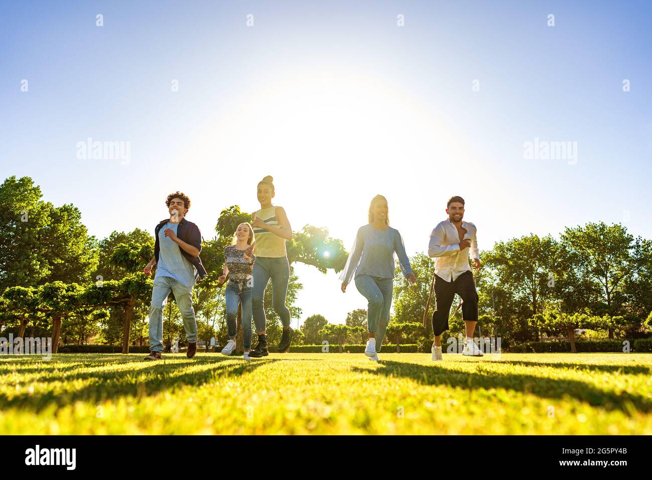 Vue sous angle suggestive d'un groupe de jeunes amis multiraciaux de gen-z qui s'exécutent sur un terrain vert avec un coucher de soleil sur leur dos avec un effet de lumière et long Banque D'Images