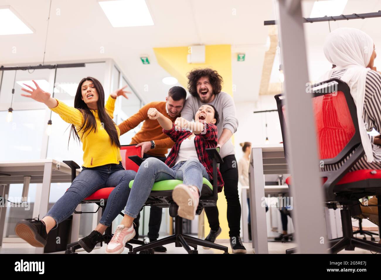 Les collègues s'amusent au travail au bureau. Filles poussant des garçons sur des chaises avec des roues. Groupe multiethnique de start-up réussie Banque D'Images