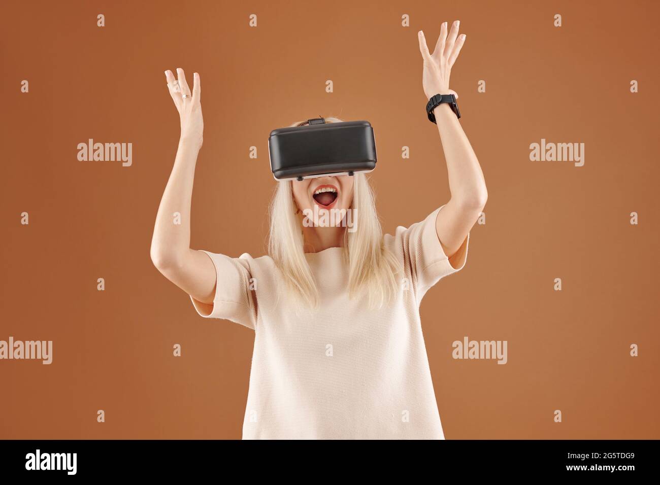 Jeune femme blonde ravie avec la bouche ouverte levant les mains dans l'excitation tout en utilisant le simulateur de réalité virtuelle sur fond marron Banque D'Images