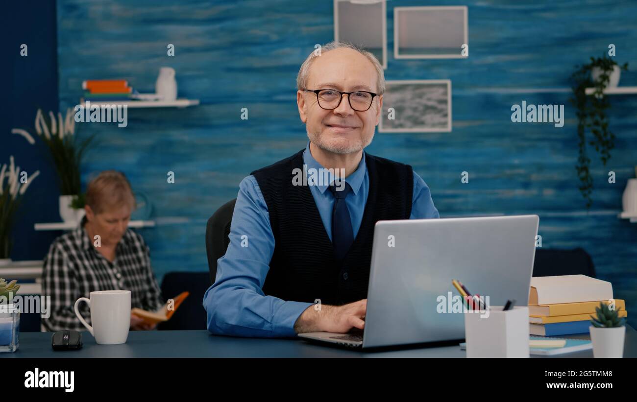 Un directeur à la retraite assis à la réception devant l'appareil photo souriant après avoir tapé sur un ordinateur portable à la maison pendant que la femme principale lisait un livre en arrière-plan. Homme préparé pour la réunion en ligne à l'aide de gadgets moden Banque D'Images
