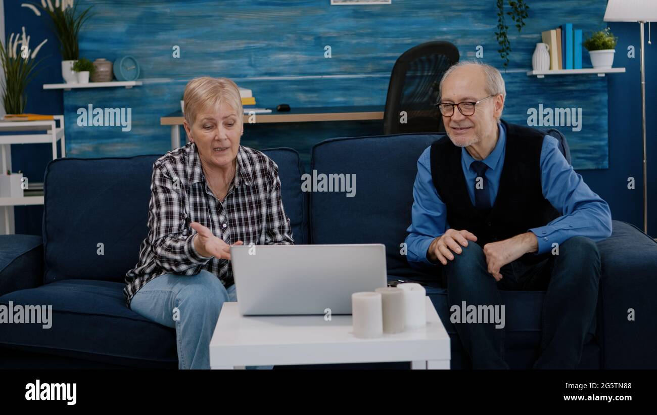 Un couple expérimenté souriant et enthousiaste regardant un ordinateur portable se faire passer pendant que le magnétoscope est assis sur un canapé. Un mari et une femme à la retraite souriant lors d'une réunion virtuelle se reposant sur un canapé dans une salle confortable. Banque D'Images