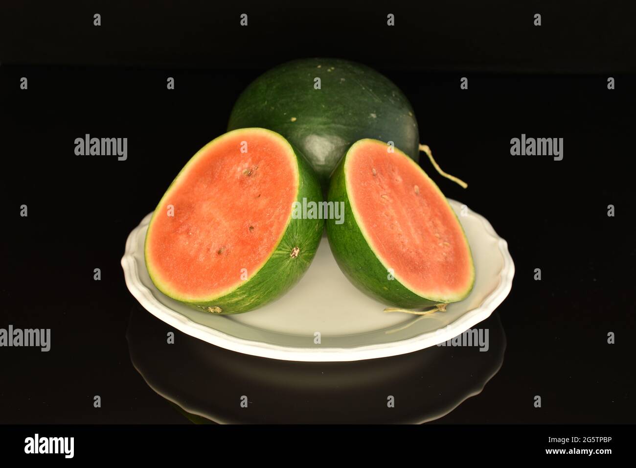 Une moitié entière et deux moitiés de pastèque biologique sur un plat blanc, gros plan, sur fond noir. Banque D'Images