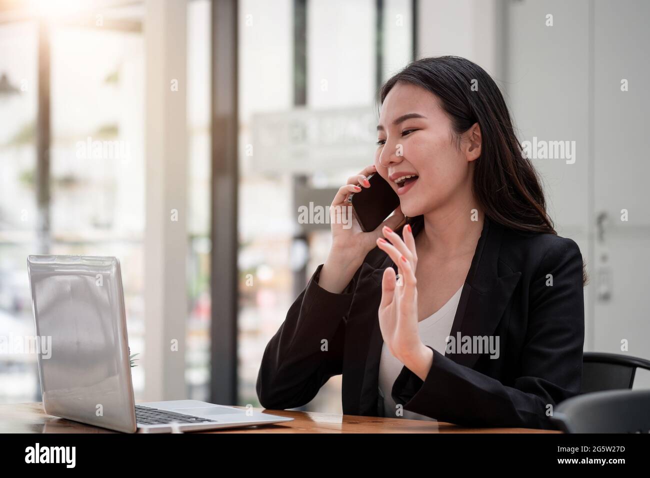 Adorable jeune femme asiatique avec grin sur le téléphone tout en travaillant sur un ordinateur portable au café-restaurant Banque D'Images