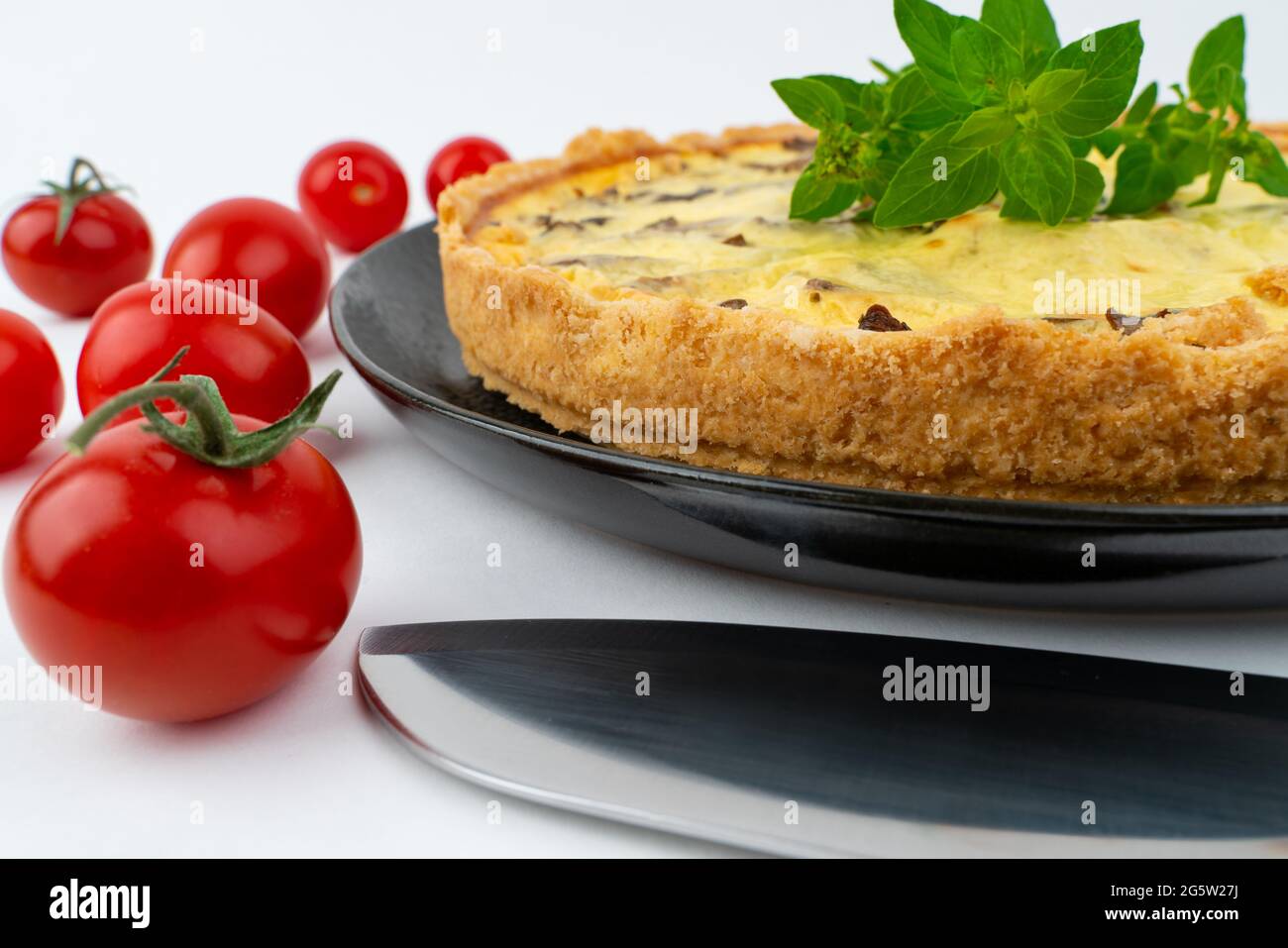 Gros plan grand studio de gâteau français salé jaune fraîchement cuit, ou quiche, avec des champignons, des tomates cerises rouges, sur une assiette design noire isolée o Banque D'Images